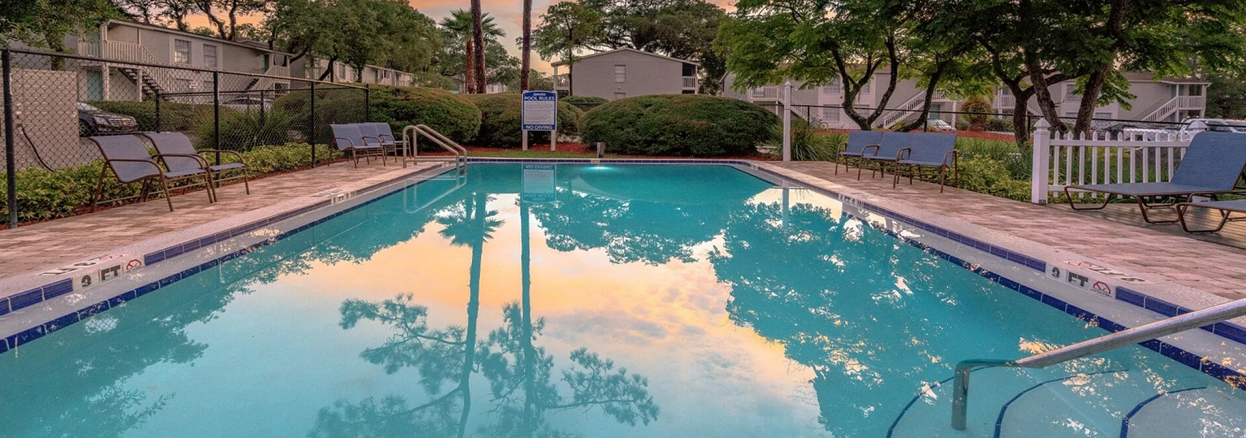 Serene apartment community pool surrounded by lush greenery and lounge chairs, set under a vibrant sunset sky.