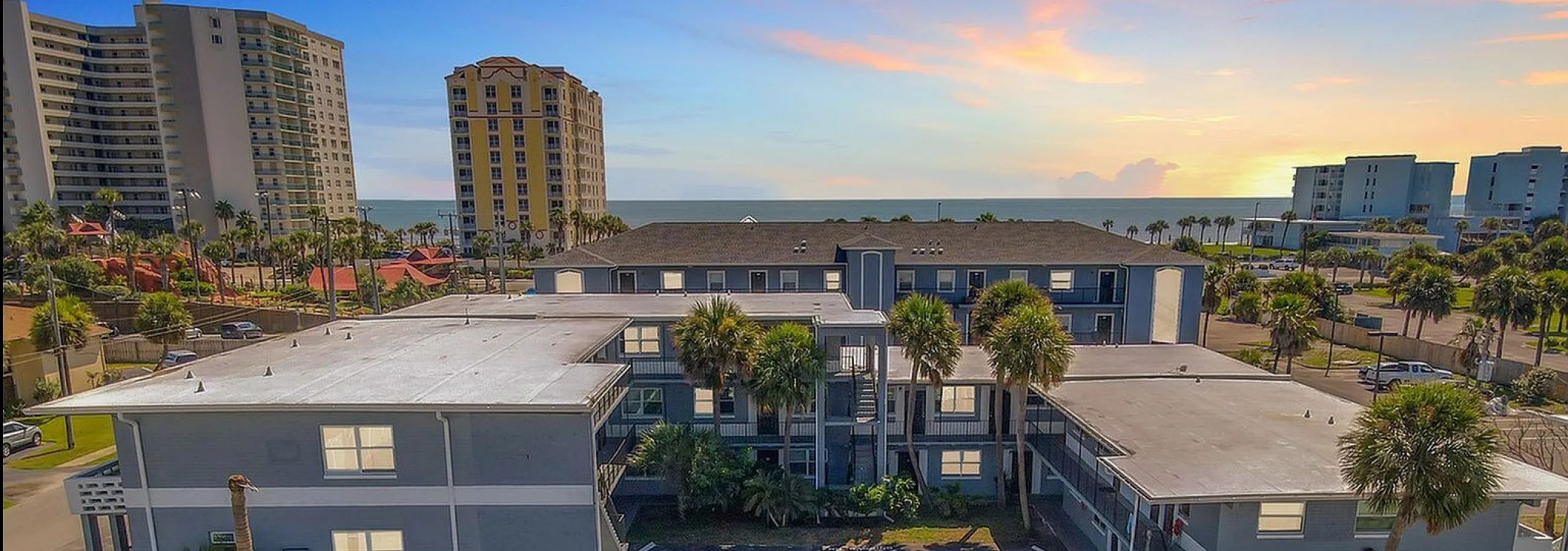 Stunning coastal apartment complex with sleek architecture, ample parking, and palm trees under a vibrant sunset sky.