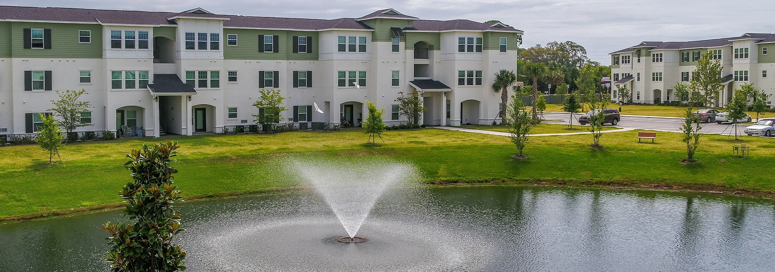 Charming apartment complex with three-story buildings overlooking a serene pond with a graceful fountain, surrounded by lush greenery.