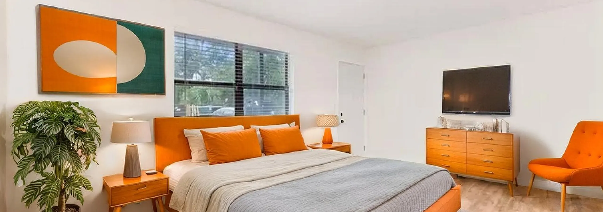 Charming bedroom with mid-century modern furniture, featuring a cozy king-sized bed with vibrant orange accents, wooden flooring, and stylish decor.