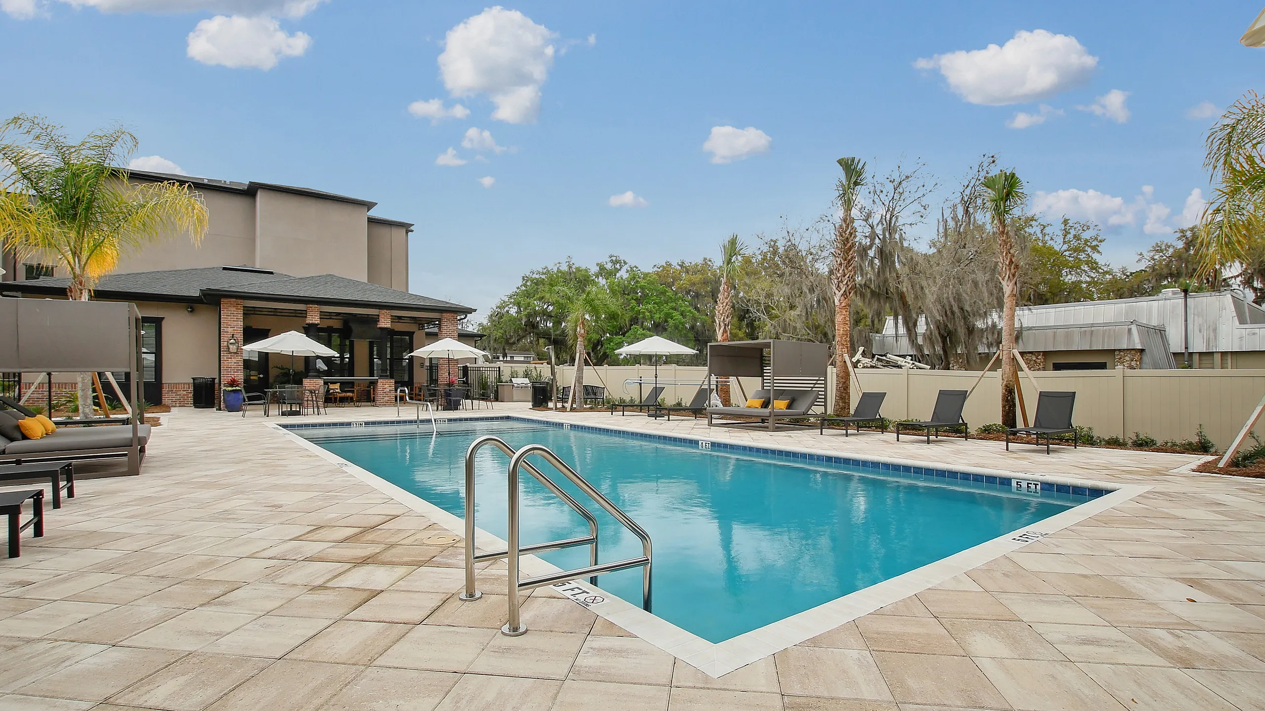 Inviting apartment community pool area with pristine blue water, surrounded by comfortable loungers, palm trees, and a modern clubhouse under a clear blue sky.