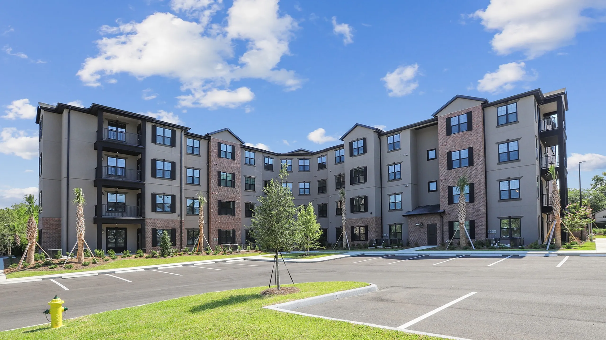Modern apartment complex with brick facade, ample parking, and lush landscaping under a bright blue sky.