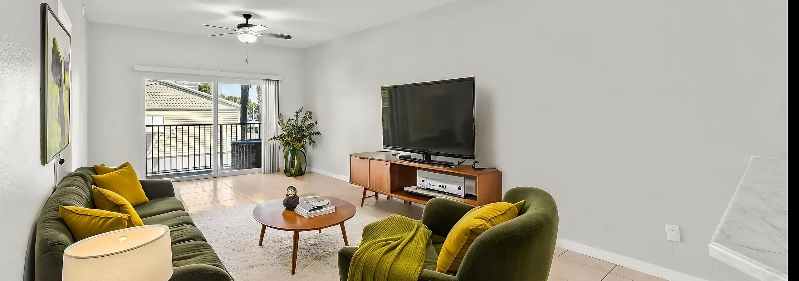 Bright and inviting living room with modern furnishings, lush green accents, and natural light from sliding glass doors opening to a balcony.