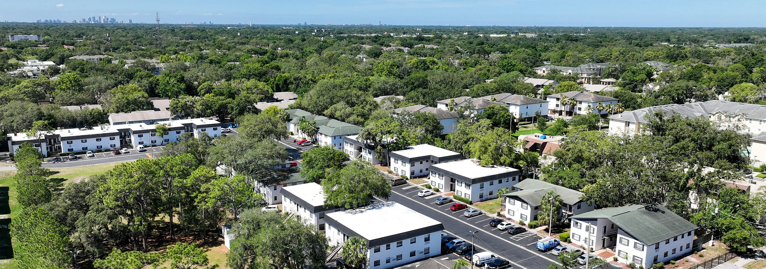 Charming aerial view of a serene suburban neighborhood with lush greenery, cozy apartment buildings, and a sparkling community pool under a clear blue sky.