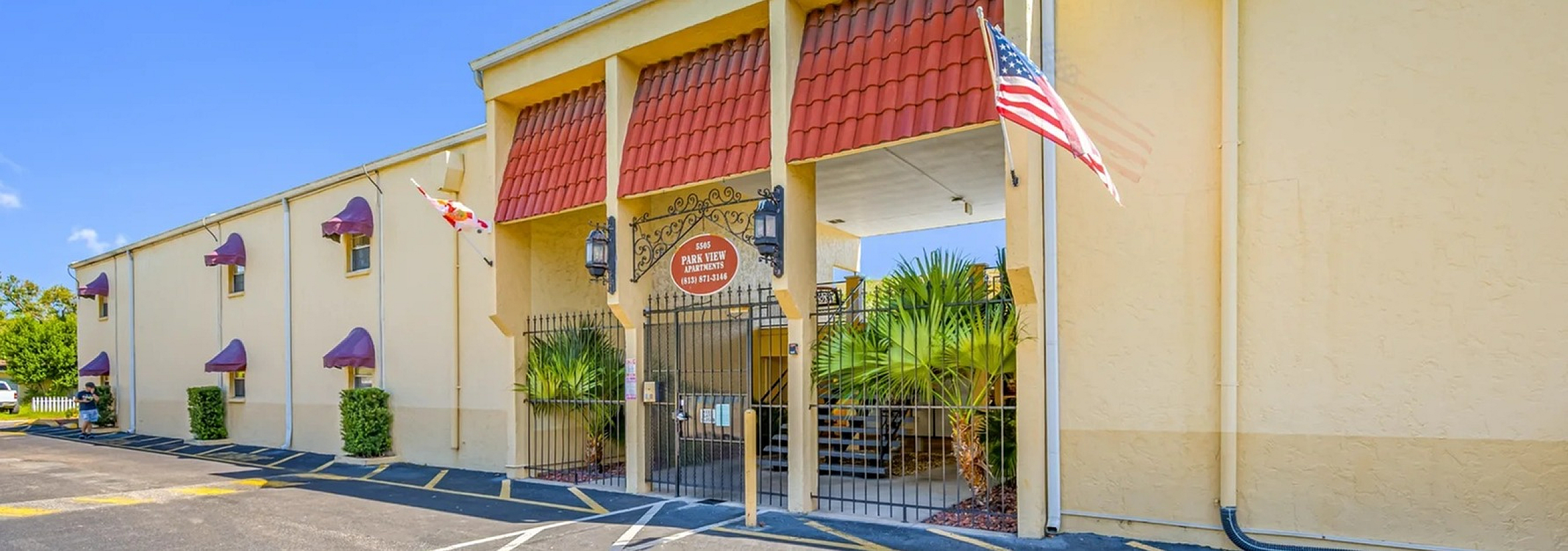 Stylish apartment building entrance with red awnings and decorative iron gate, surrounded by lush palm landscaping.