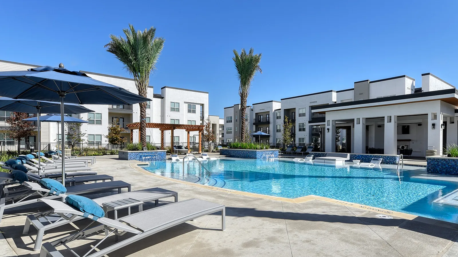 Luxurious apartment complex featuring a sparkling pool surrounded by stylish lounge chairs and elegant palm trees under a clear blue sky.