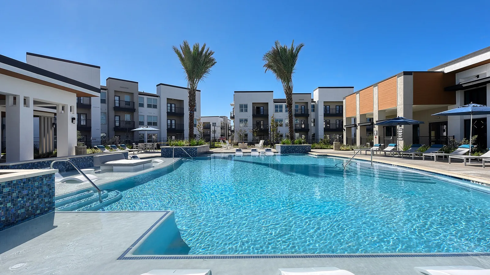 Luxurious apartment complex featuring a stunning outdoor pool with modern lounge areas and palm trees under a clear blue sky.