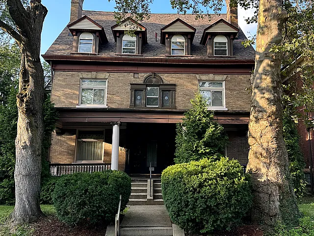 Charming three-story brick building with classic dormer windows, nestled between lush greenery and mature trees.