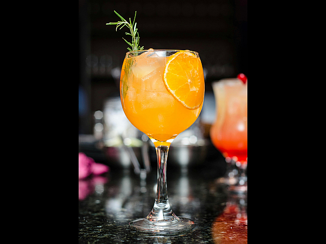 Vibrant orange cocktail garnished with fresh rosemary and a slice of orange, elegantly served in a wine glass on a sleek black counter.