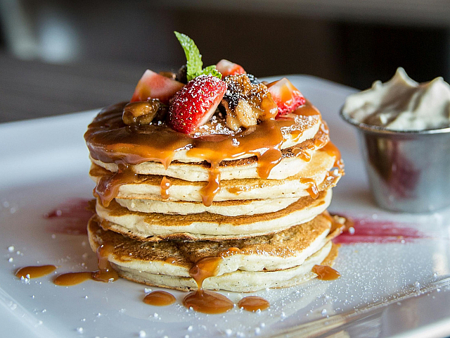 Delectable stack of pancakes drizzled with syrup and topped with fresh strawberries and walnuts, served with a side of whipped cream.