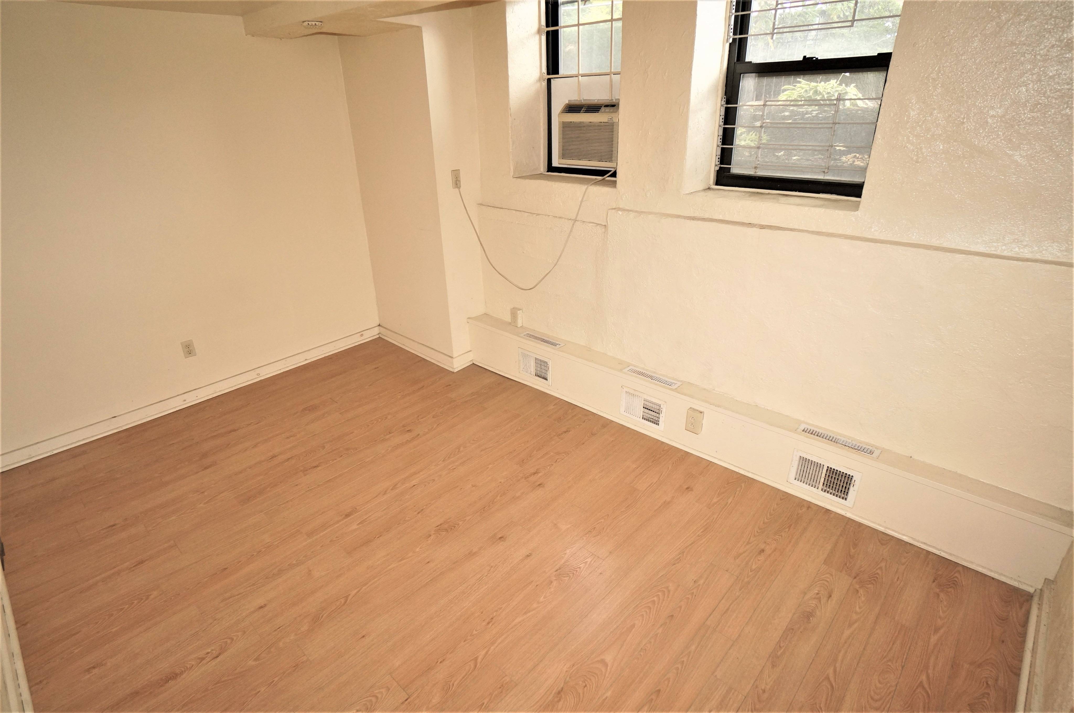 Bedroom angle shows windows, and air conditioner. Laminate floors and white walls throughout.