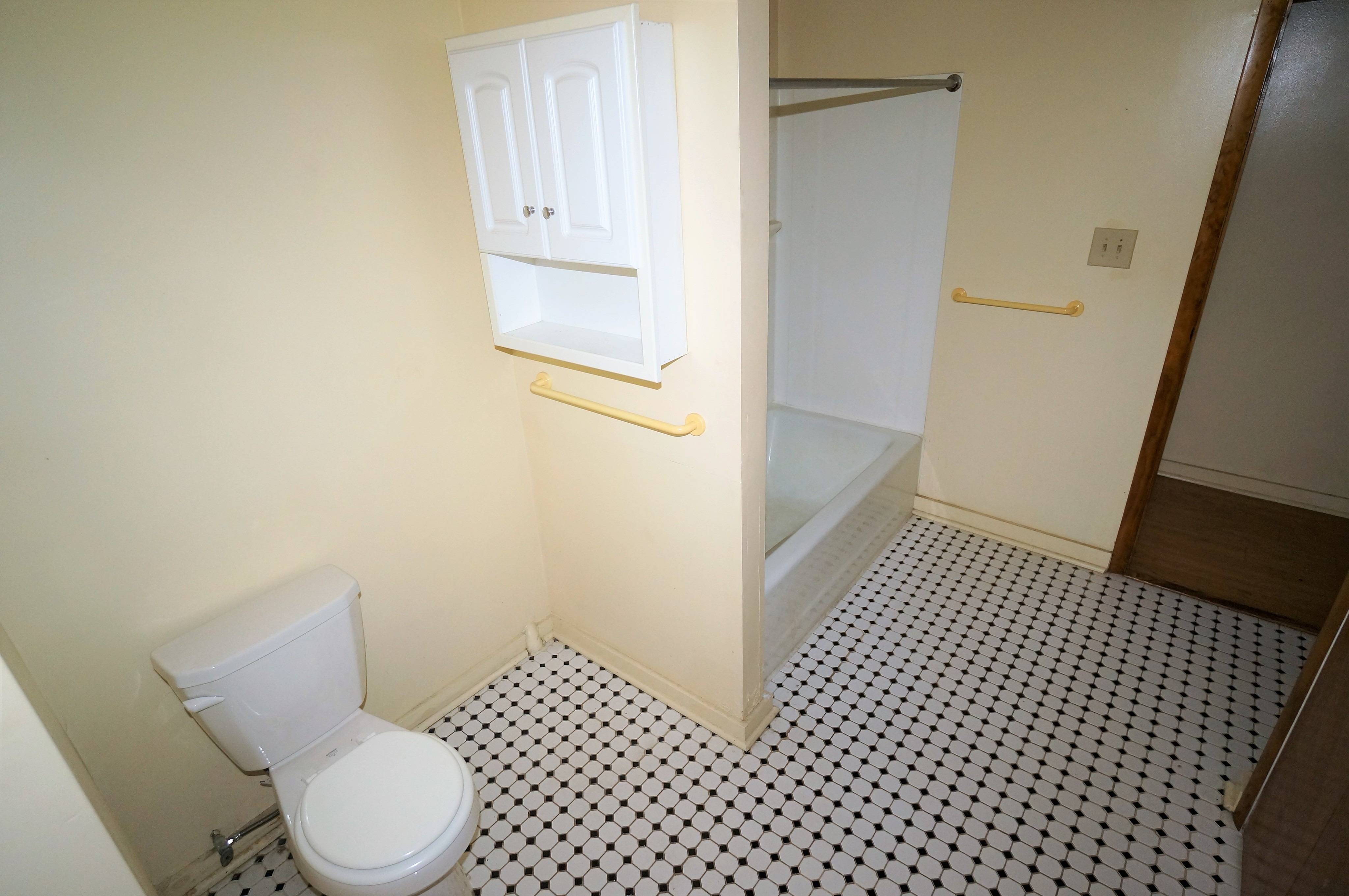 bathroom shown has white walls with white tile floors. there is a white sink, toilet, and tub, with a wooden vanity and accompanying mirror.