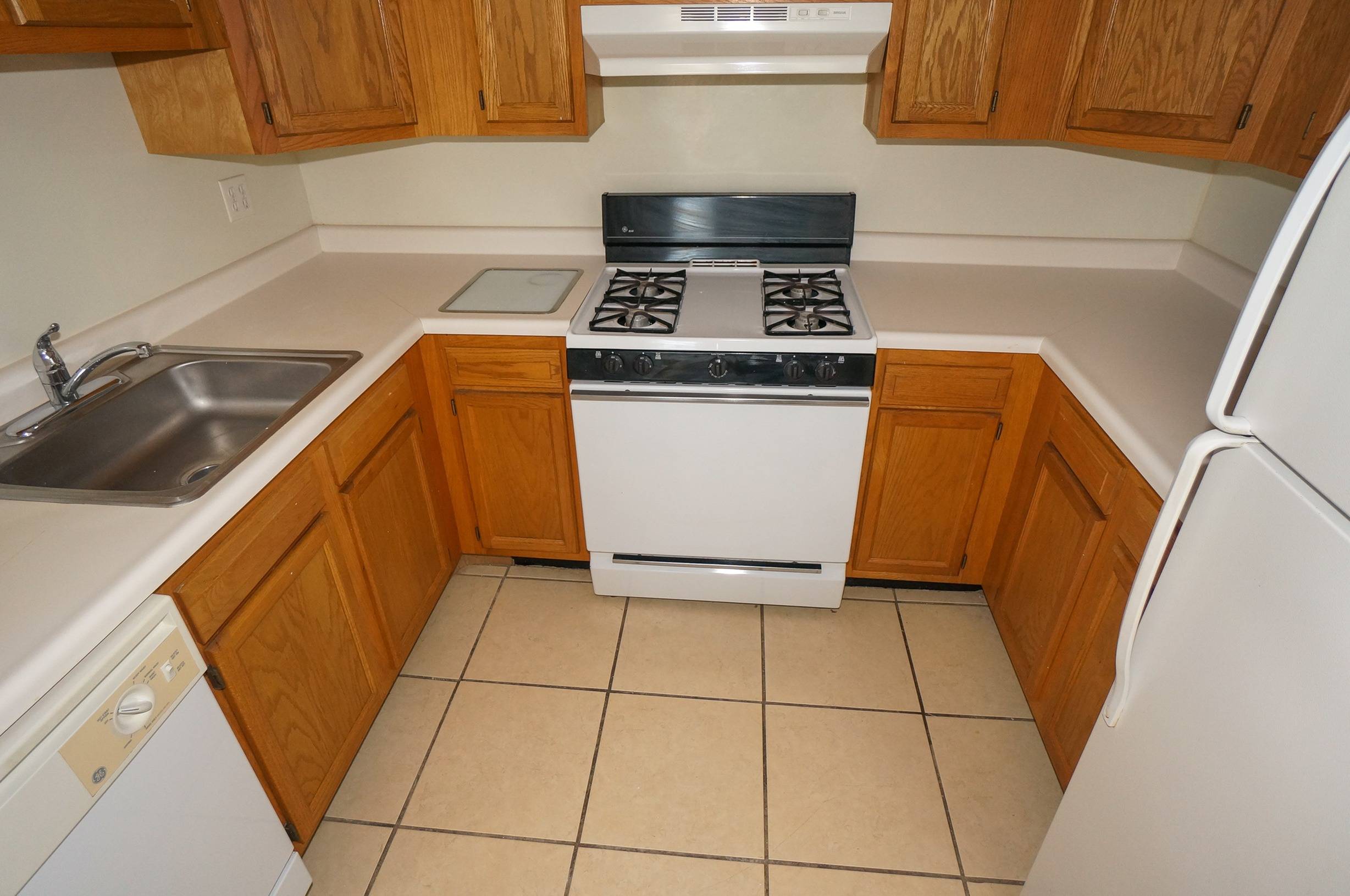 Kitchen shown has tile flooring, with wood cabinets, Light counters, as well as a white fridge, and white gas oven/stove combination and a dishwasher
