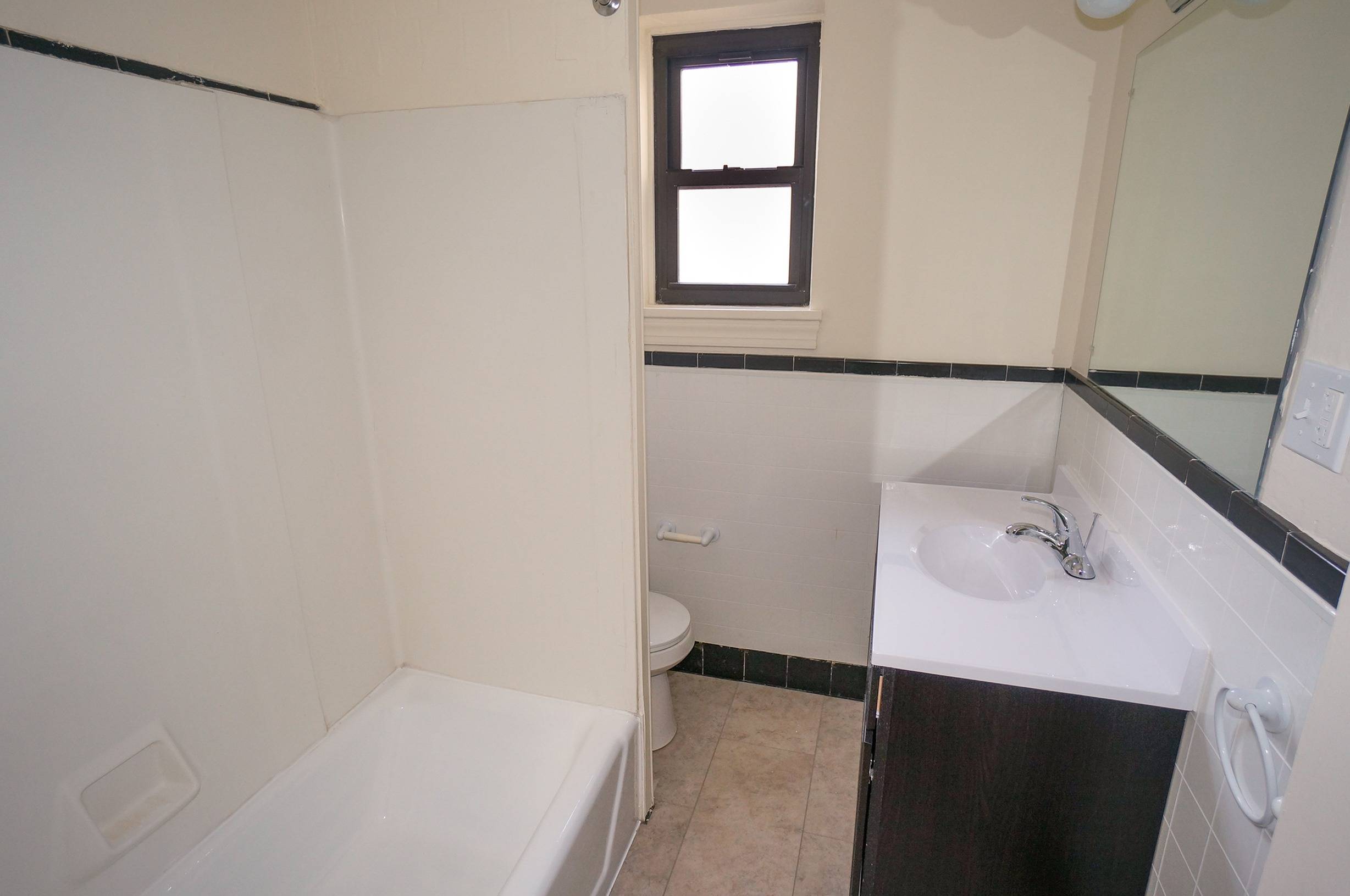 bathroom shown has white tile walls with white tile floors. there is a white sink, toilet, and tub, with a wooden vanity and accompanying mirror.
