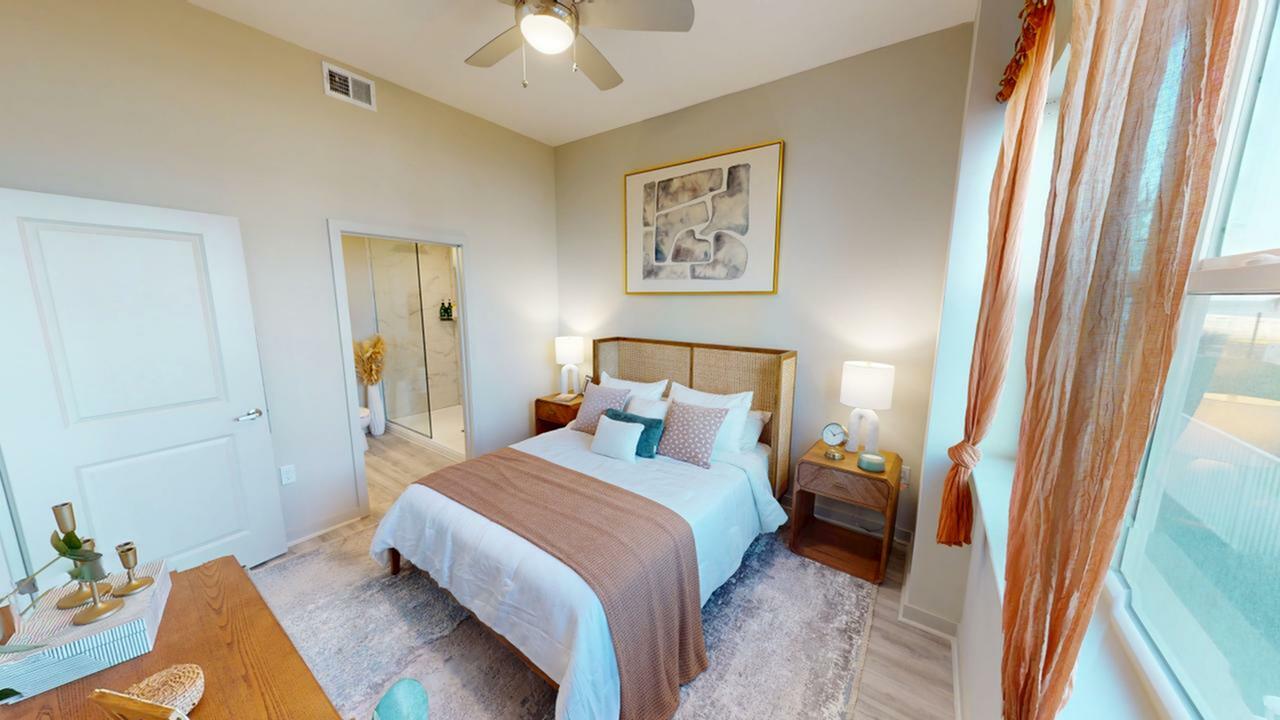 Inviting bedroom with elegant decor, featuring a cozy bed with plush pillows, modern bedside tables, ambient lighting, and a stylish artwork. The room is brightened by natural light streaming through large windows with sheer curtains.