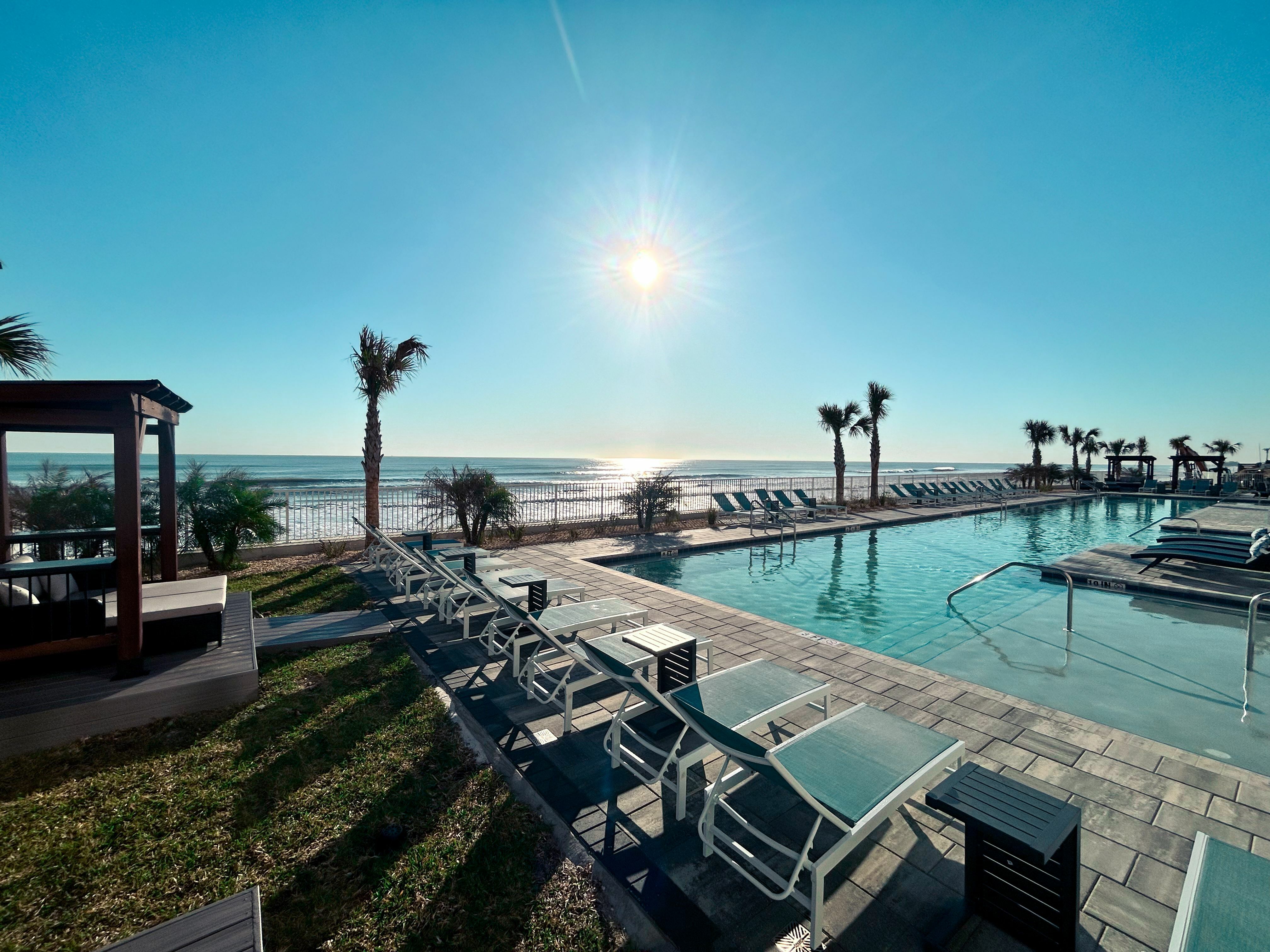 Luxurious beachfront pool area with sun loungers, palm trees, and stunning ocean views under a bright blue sky.
