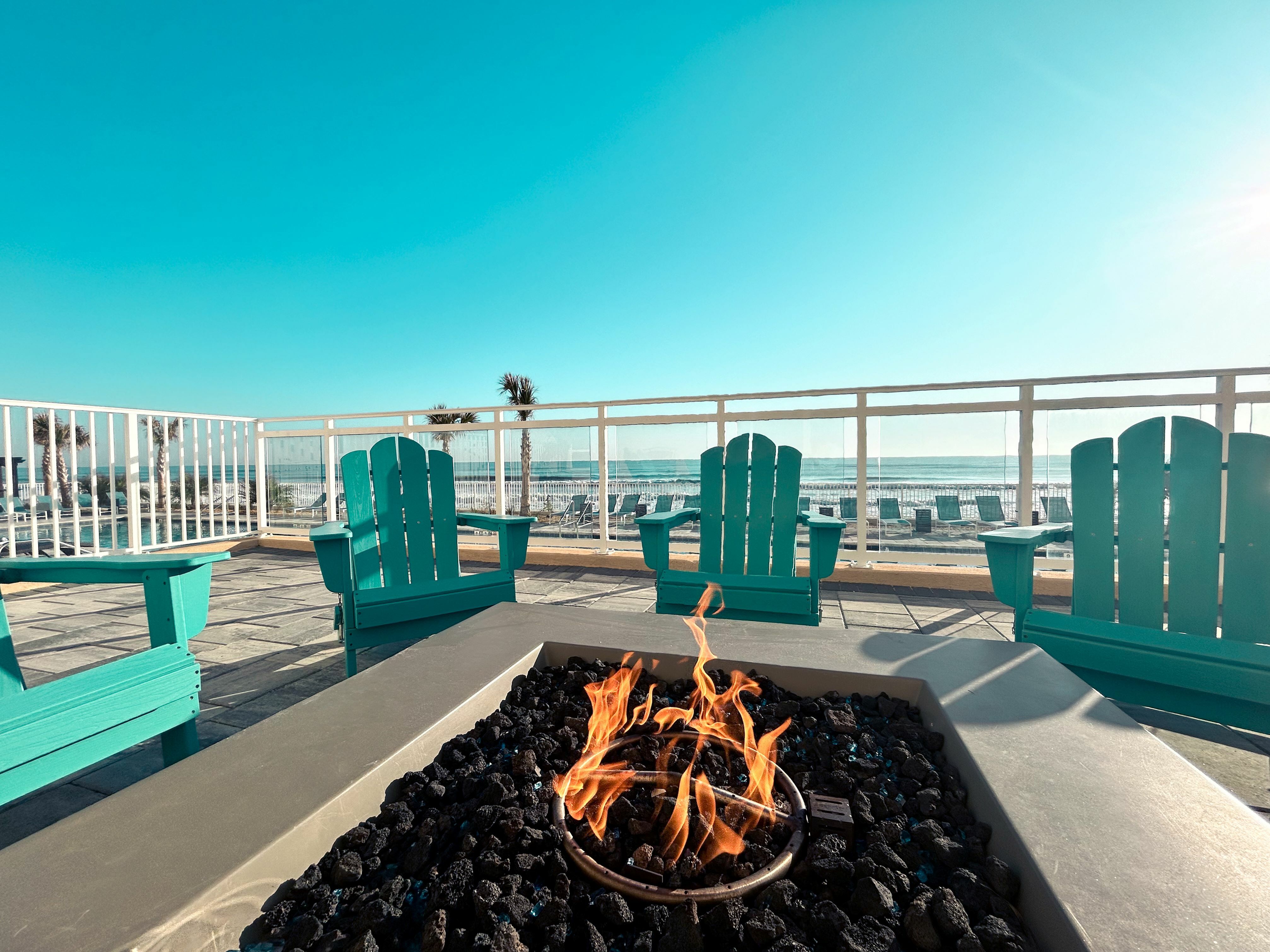 Inviting beachfront patio with turquoise Adirondack chairs around a cozy fire pit, offering panoramic ocean views.