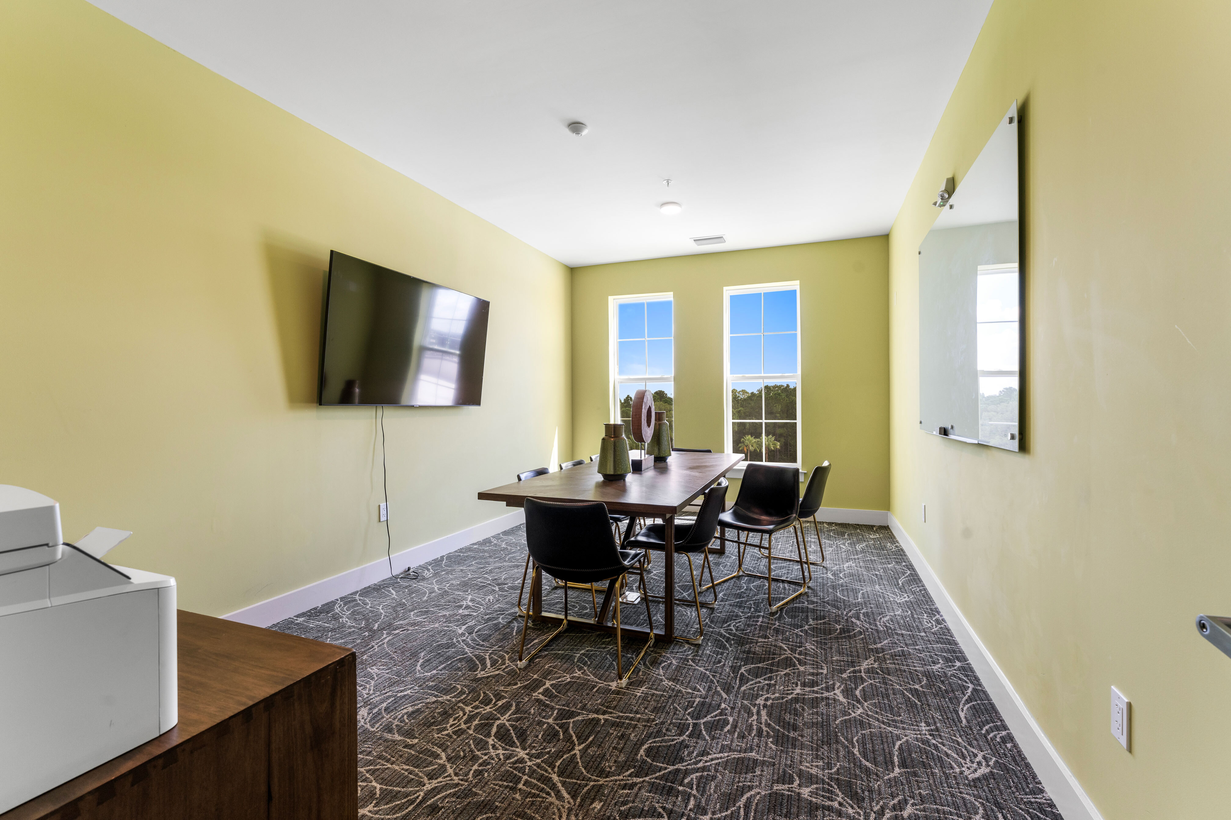 Modern conference room with vibrant yellow walls, large windows offering natural light, sleek table, comfortable chairs, and mounted flat-screen TV.