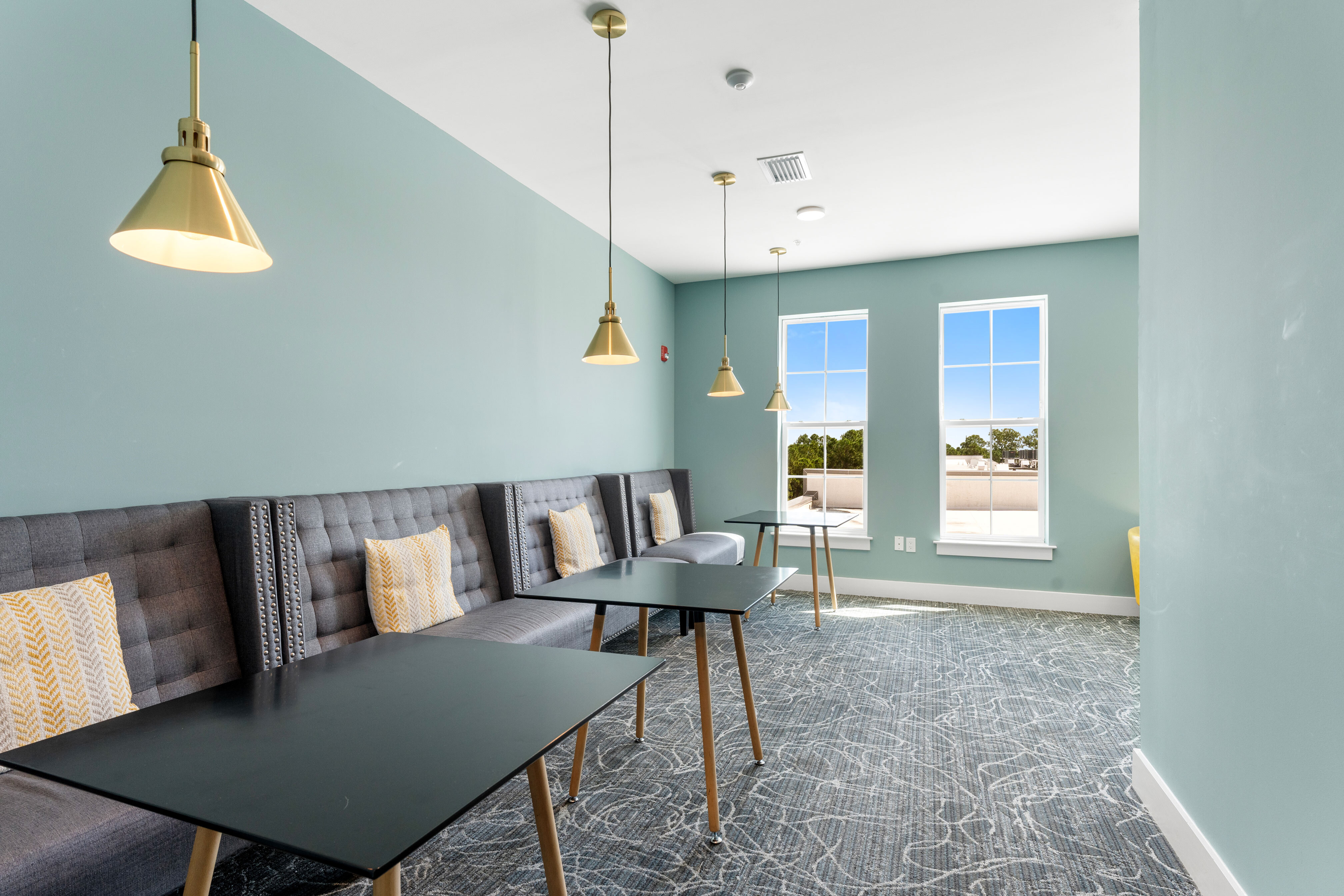 Modern lounge area featuring elegant pendant lighting, stylish gray seating with accent cushions, and abundant natural light from large windows.