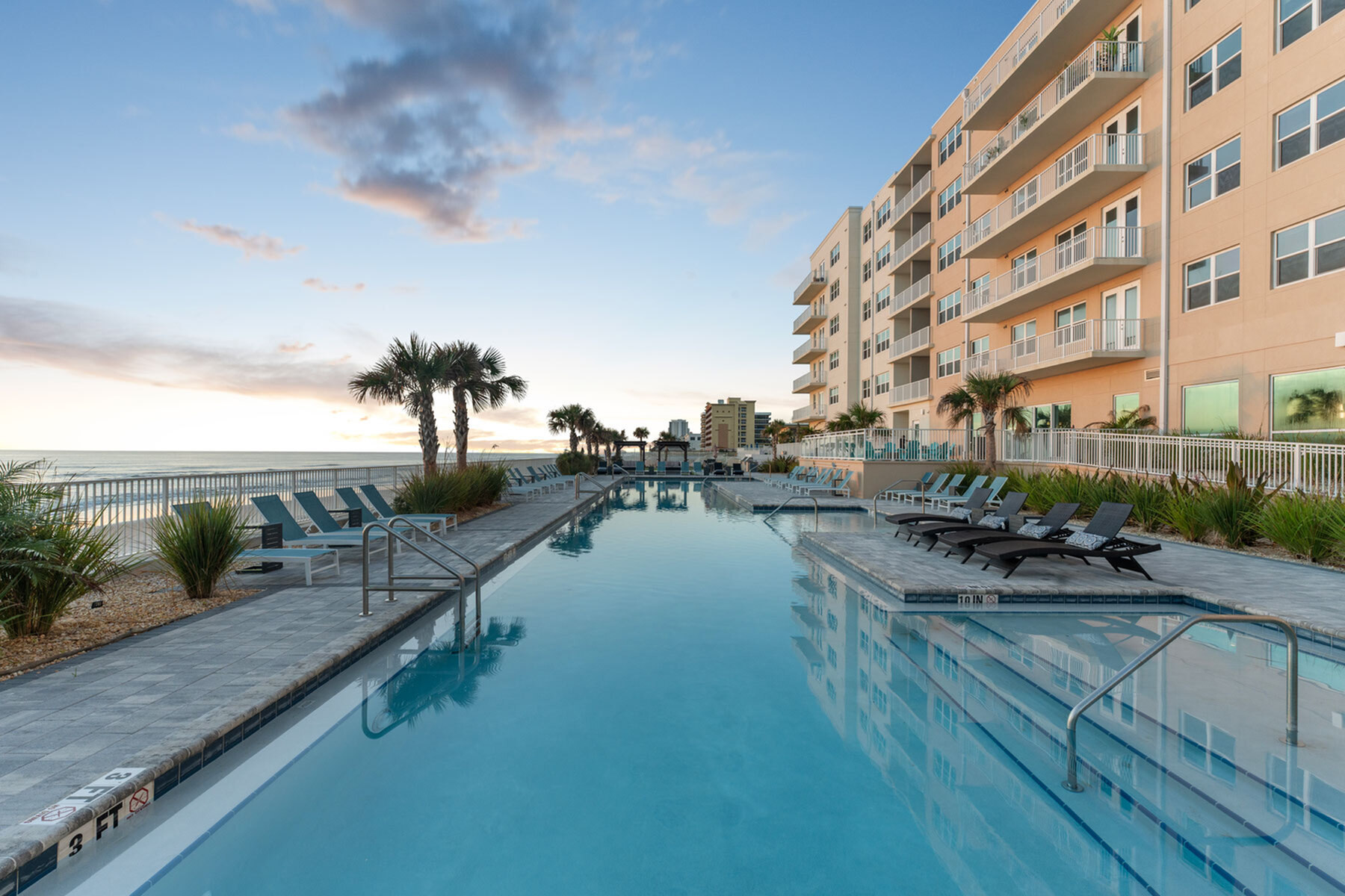 Luxurious beachfront apartment complex featuring a stunning infinity pool with ocean views, surrounded by palm trees and elegant lounge chairs.
