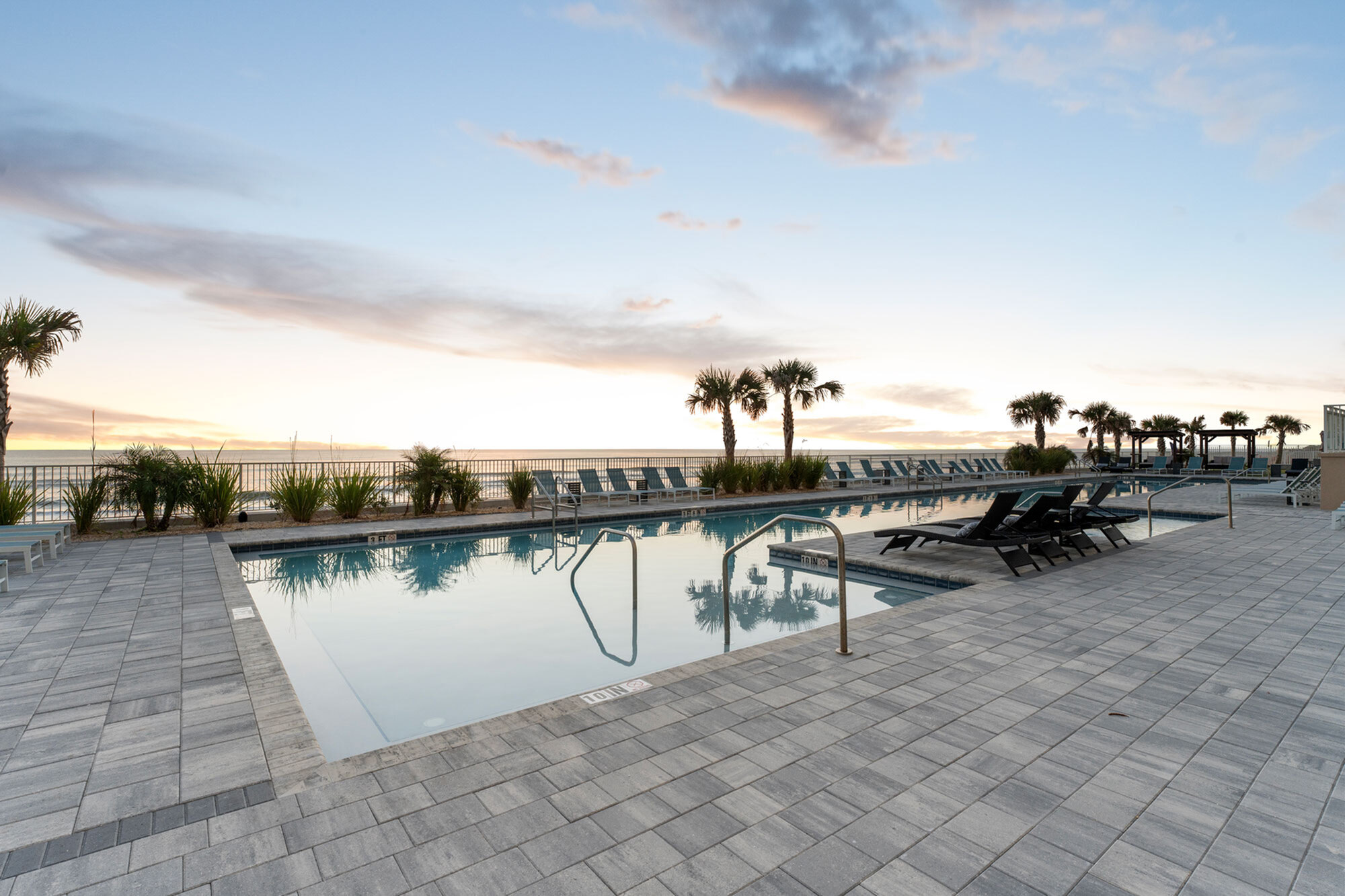 Luxurious waterfront pool area with palm trees, sun loungers, and a stunning ocean view at sunset.
