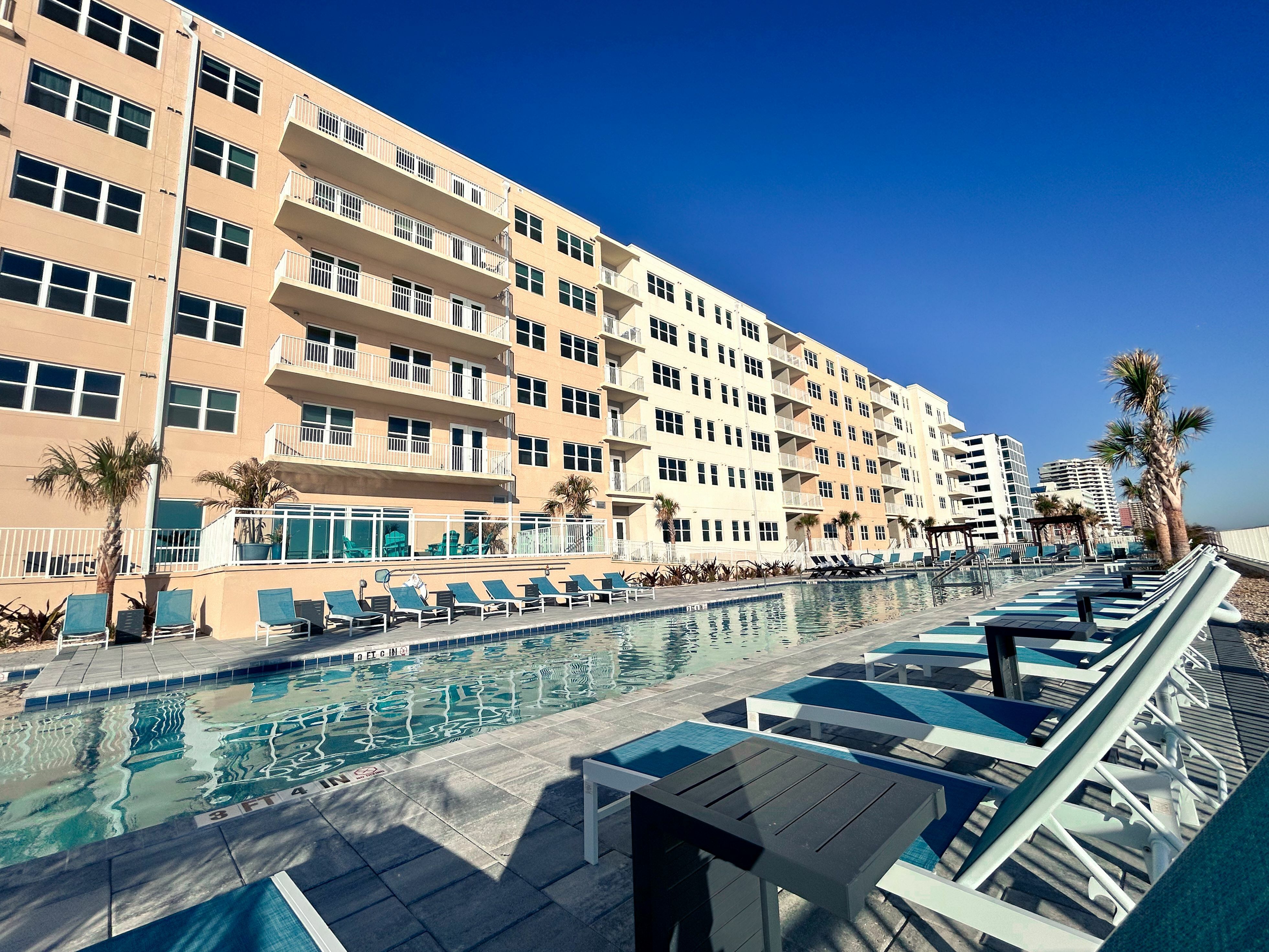 Luxurious beachfront apartment complex with stunning poolside views, modern balconies, and palm-lined surroundings under a clear blue sky.