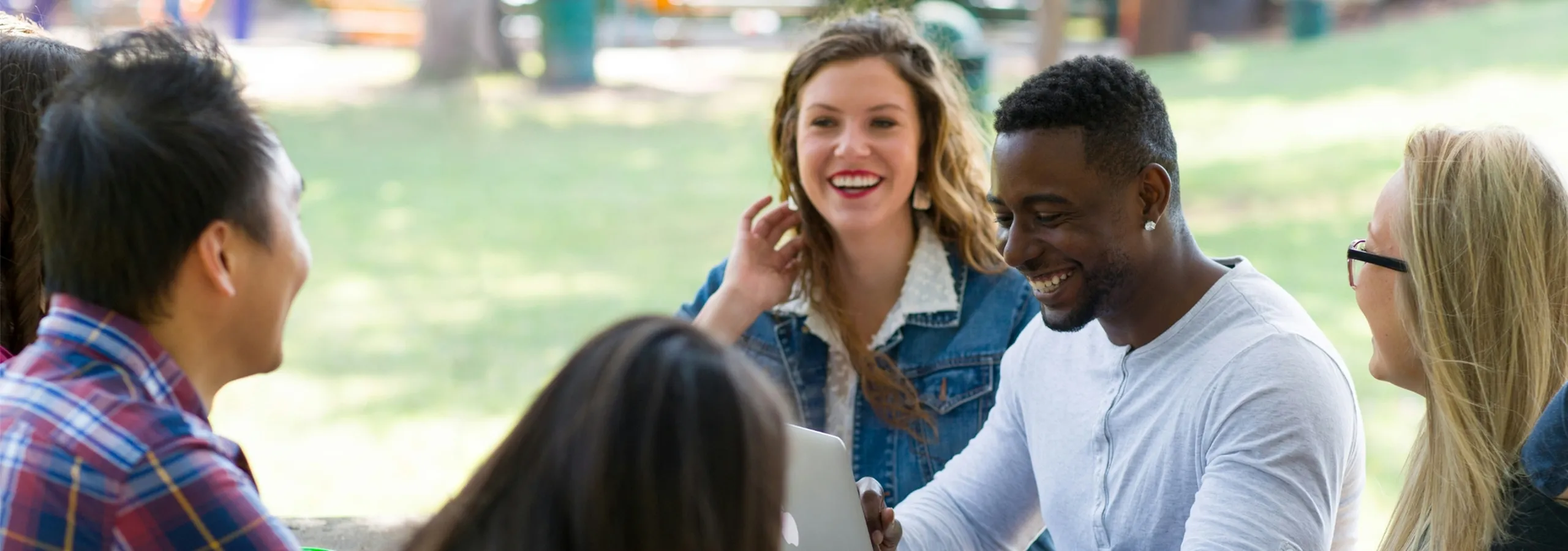 Group of friends enjoying a sunny day outdoors, sharing smiles and laughter while gathered around a laptop.