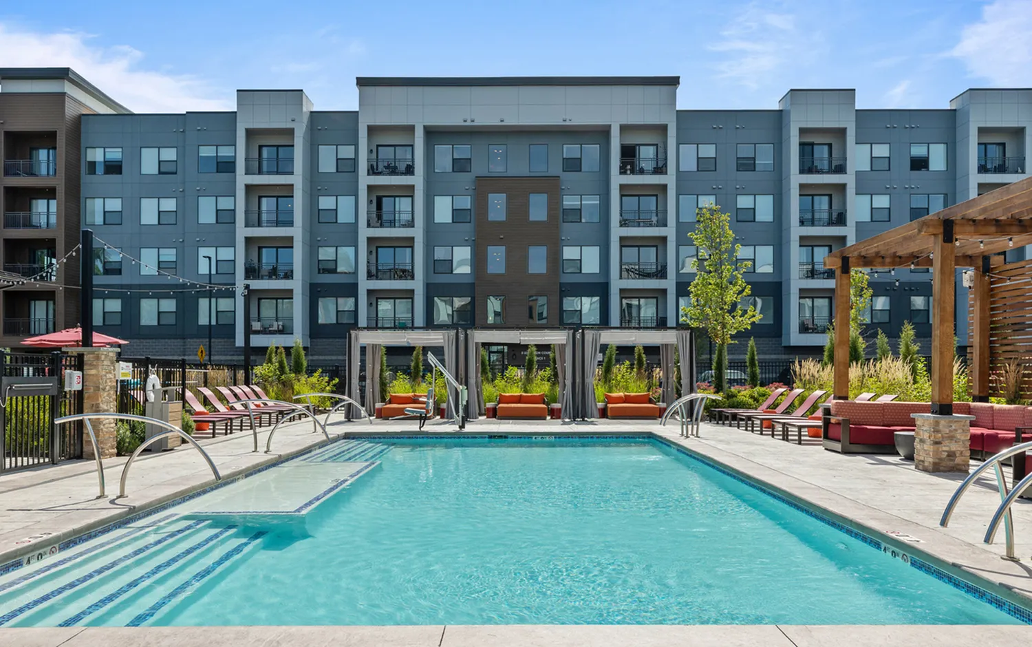 Vibrant pool area in front of a modern apartment complex with poolside seating and greenery.