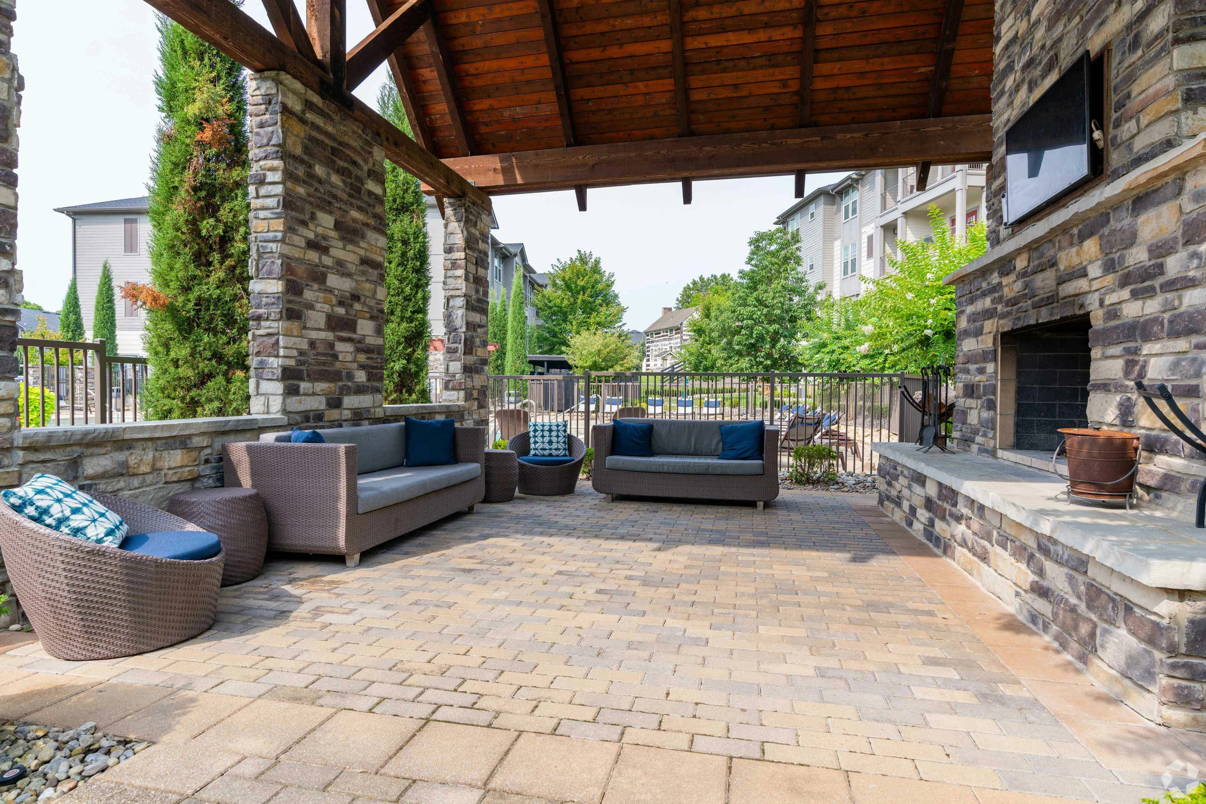 Inviting outdoor lounge with wicker sofas under a wooden pergola, complemented by a stone fireplace and lush surroundings.