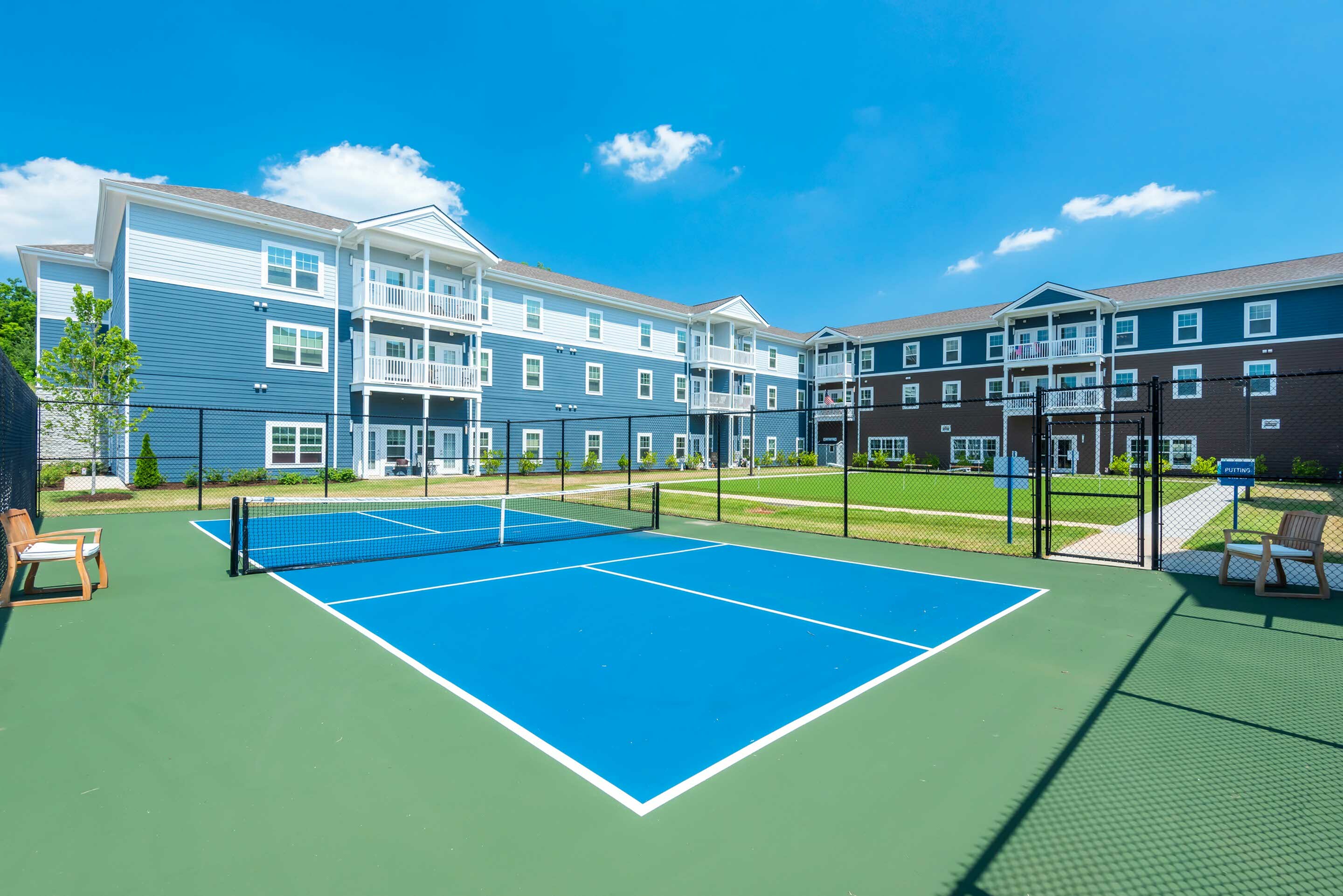 Bright and modern apartment complex with charming balconies overlooking a vibrant, blue pickleball court, set against a clear blue sky.