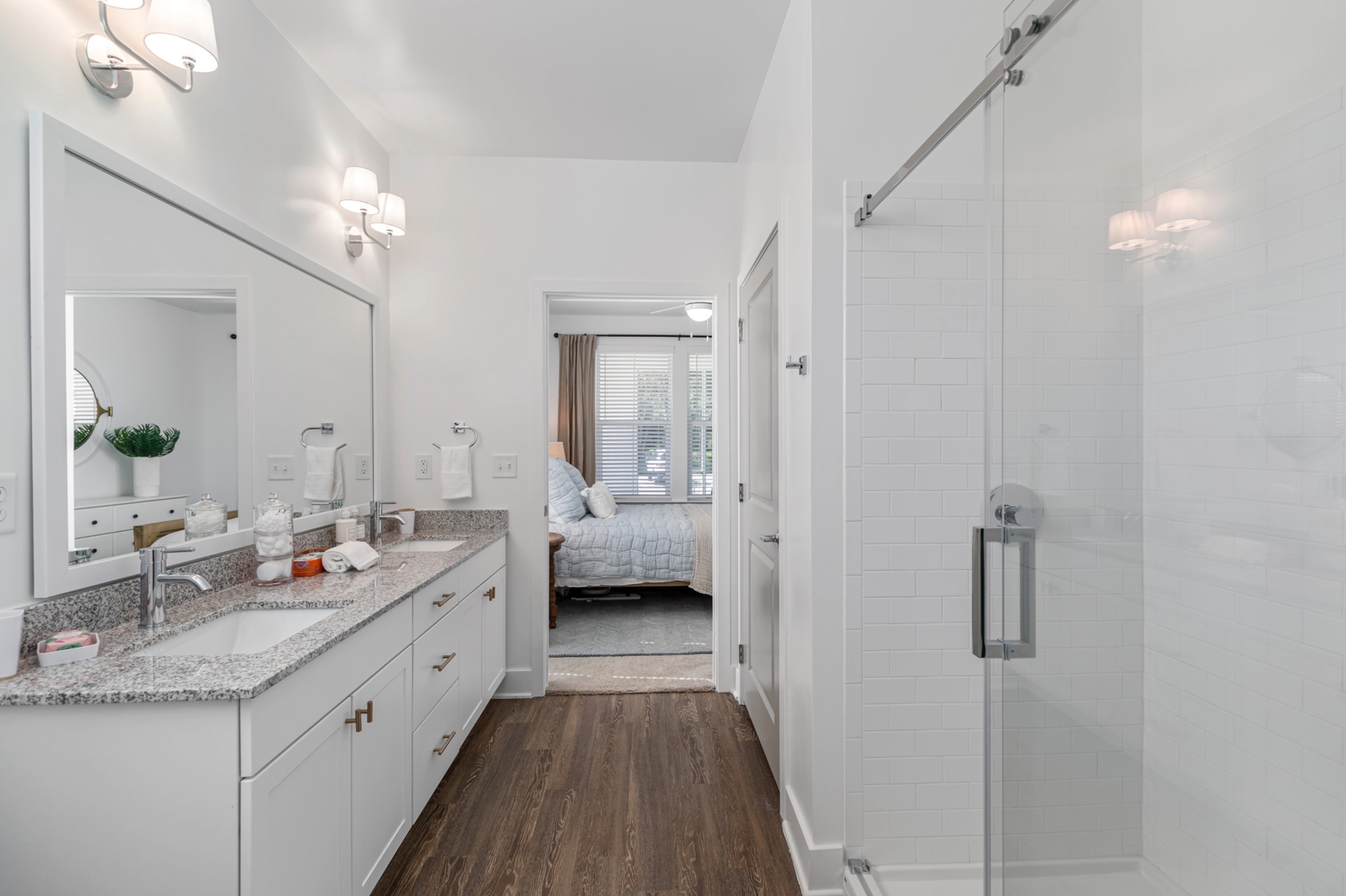 Elegant bathroom with granite double sink, sleek shower, and modern lighting leading to a cozy bedroom with natural light.