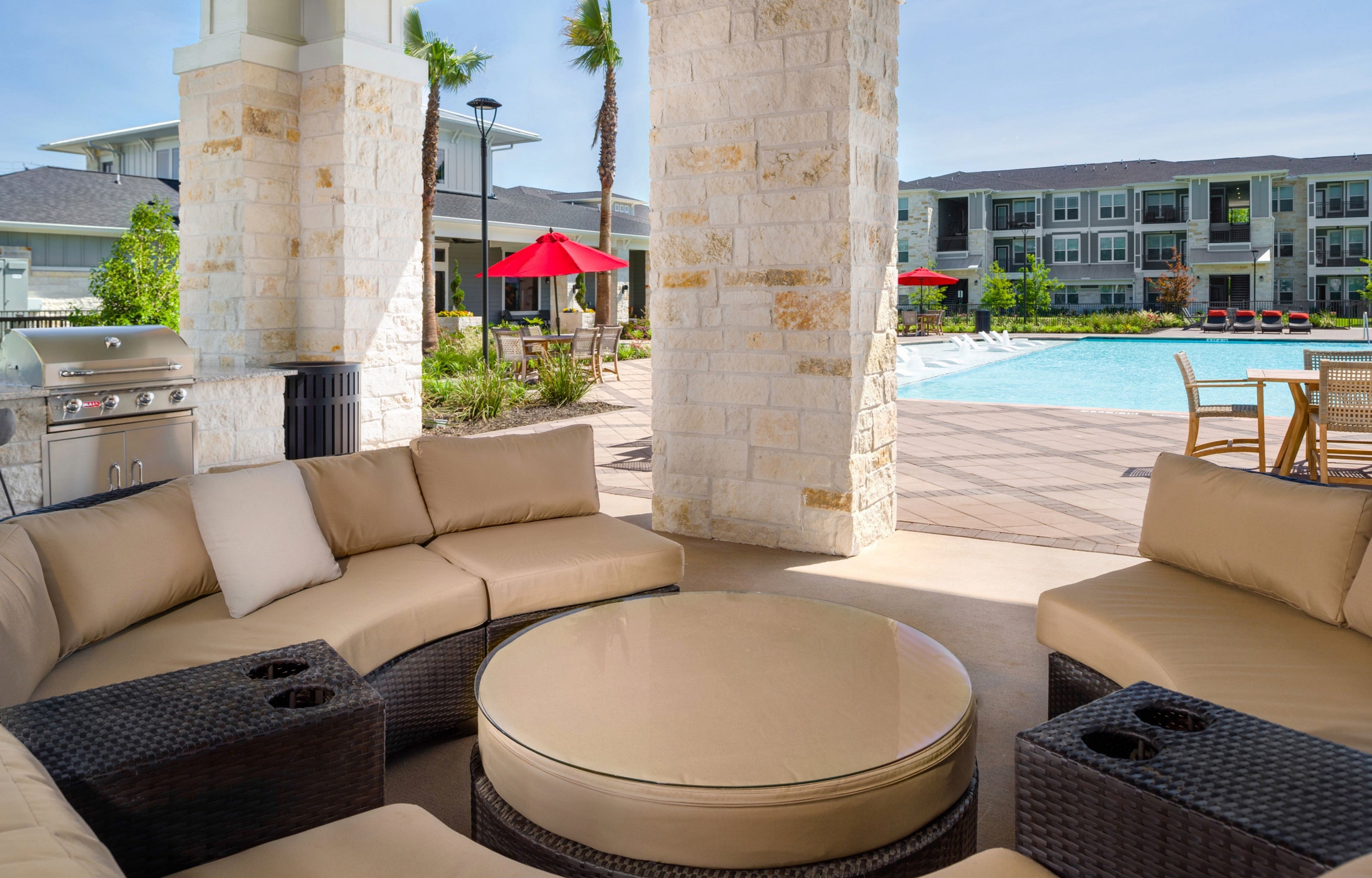Elegant outdoor seating area with plush sofas and a modern grill, overlooking a sparkling pool surrounded by red umbrellas and lush greenery.
