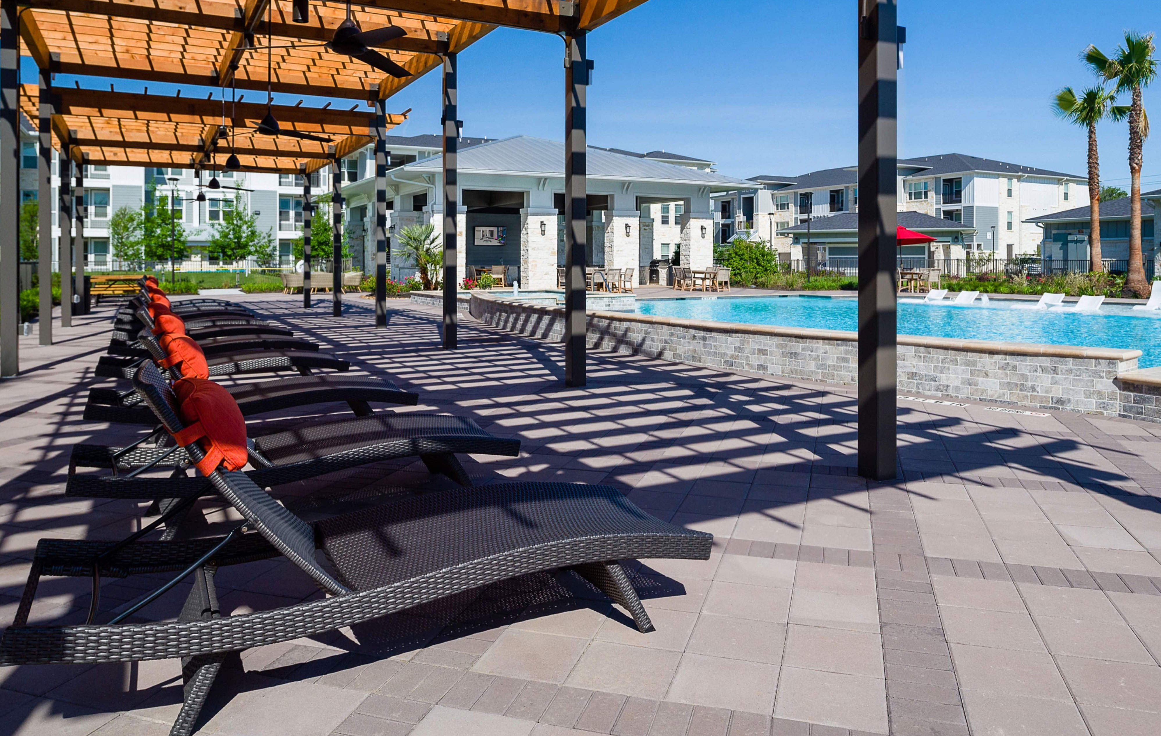 Luxury apartment complex with inviting poolside lounge chairs under a stylish pergola, surrounded by lush landscape and modern architecture.