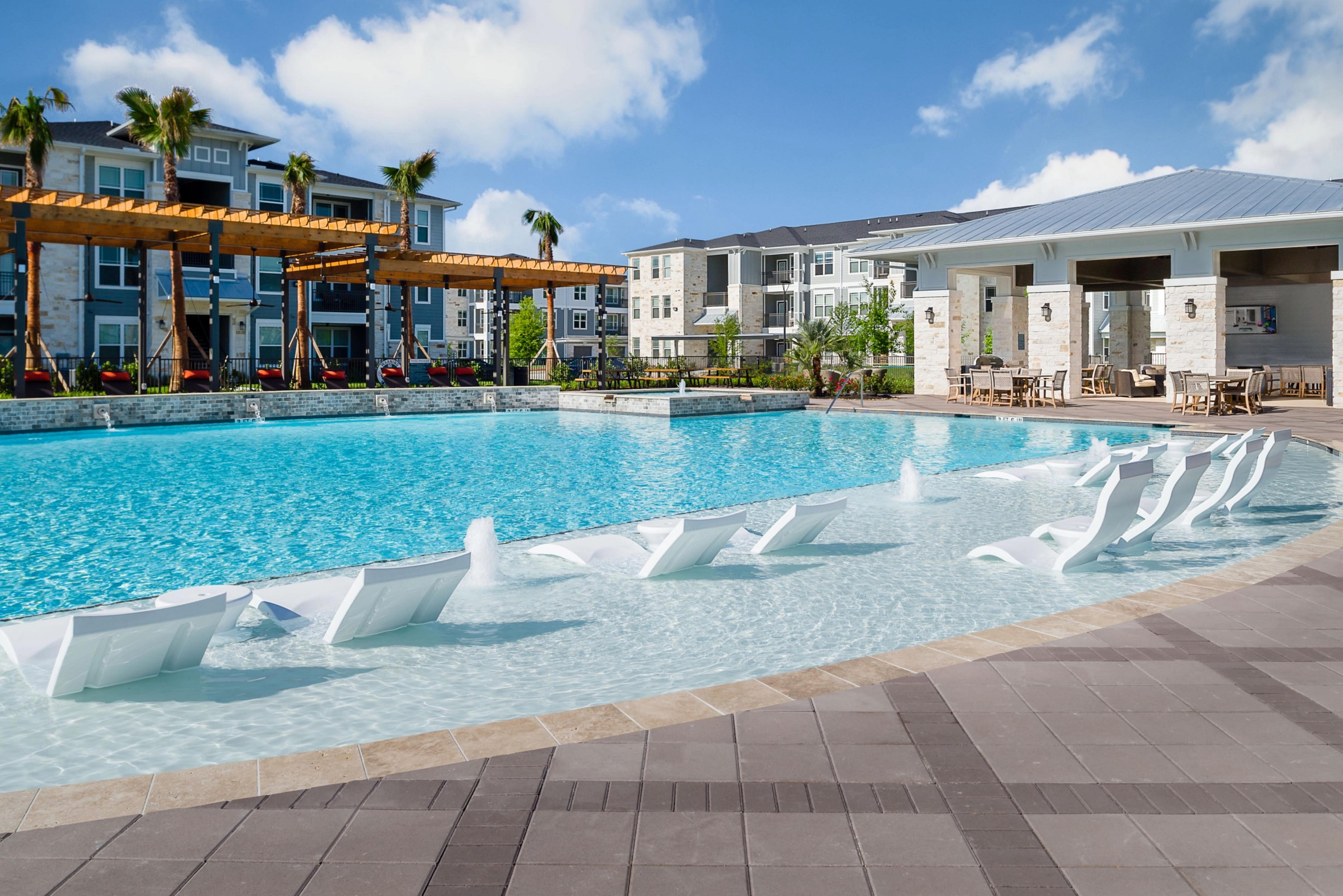 Luxurious apartment complex with resort-style pool, modern lounge chairs, and sunlit patio under clear blue skies.