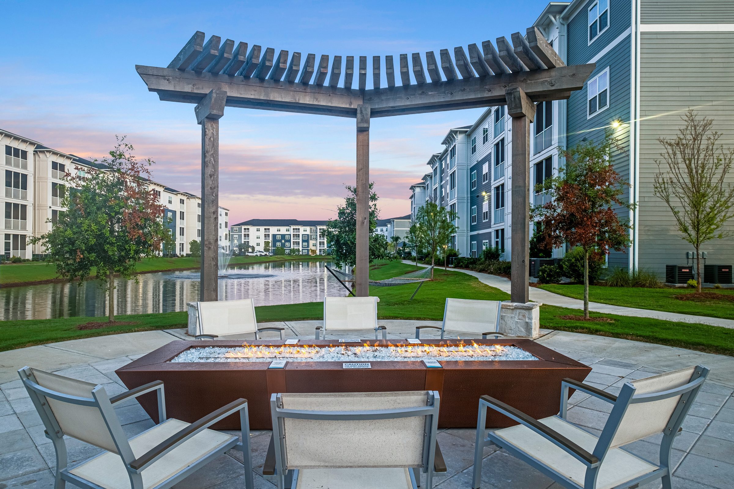 Inviting outdoor seating area with modern fire pit, framed by elegant pergola and surrounded by picturesque waterfront views of stylish apartment buildings at sunset.