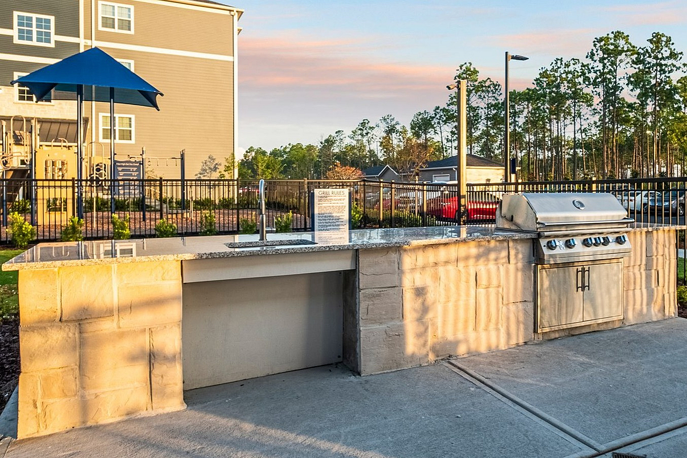 Modern apartment complex with a stylish outdoor grilling area and fenced playground, surrounded by lush greenery and scenic views.