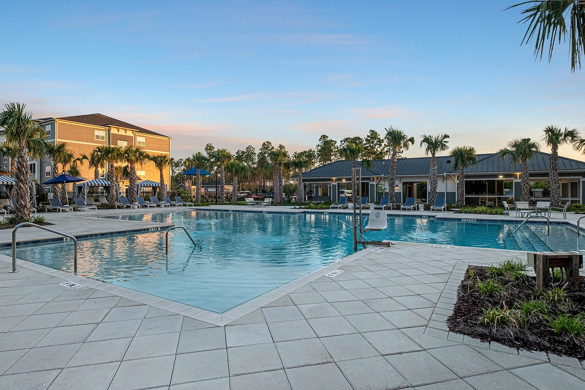 Luxurious apartment complex with a scenic resort-style swimming pool, surrounded by palm trees and elegant lounge chairs at sunset.