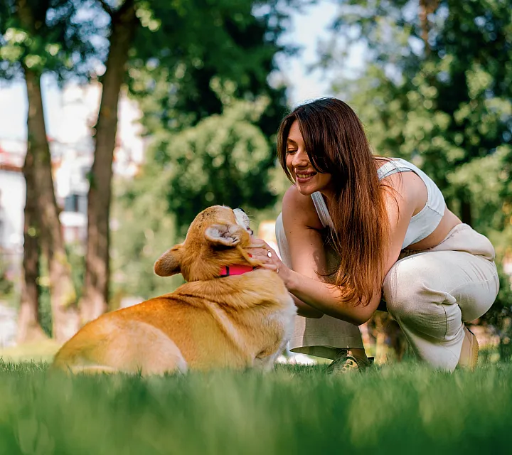 A cheerful woman enjoying a sunny day in the park with her corgi on lush green grass.