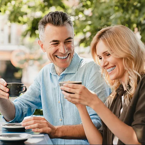 A joyful couple enjoying coffee at an outdoor café on a sunny day, surrounded by greenery.
