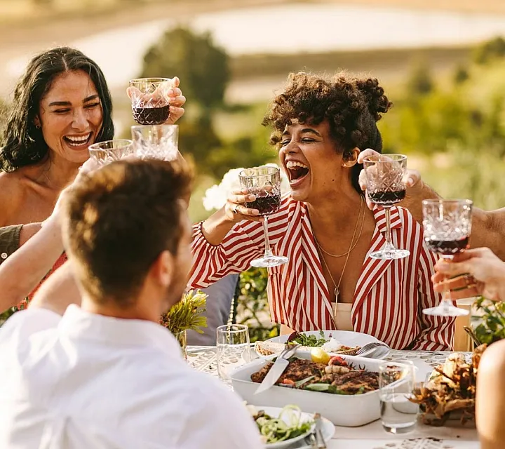 Group of friends enjoying an outdoor meal with wine, laughter, and vibrant scenery in the background.