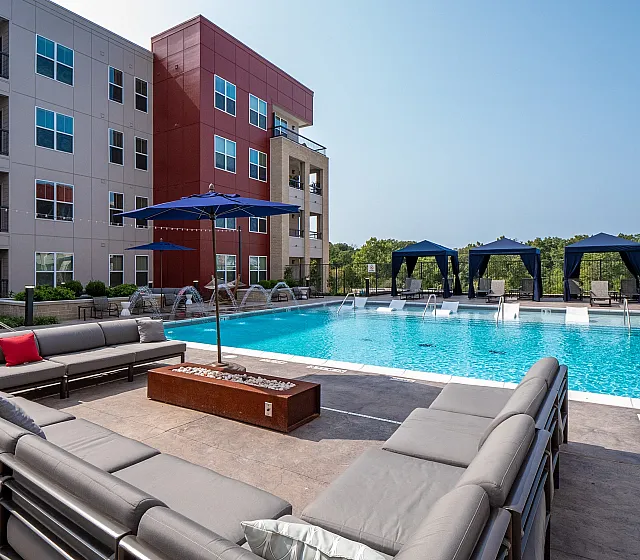 Modern apartment complex with a luxurious pool area featuring lounge seating and cabanas under a clear blue sky.