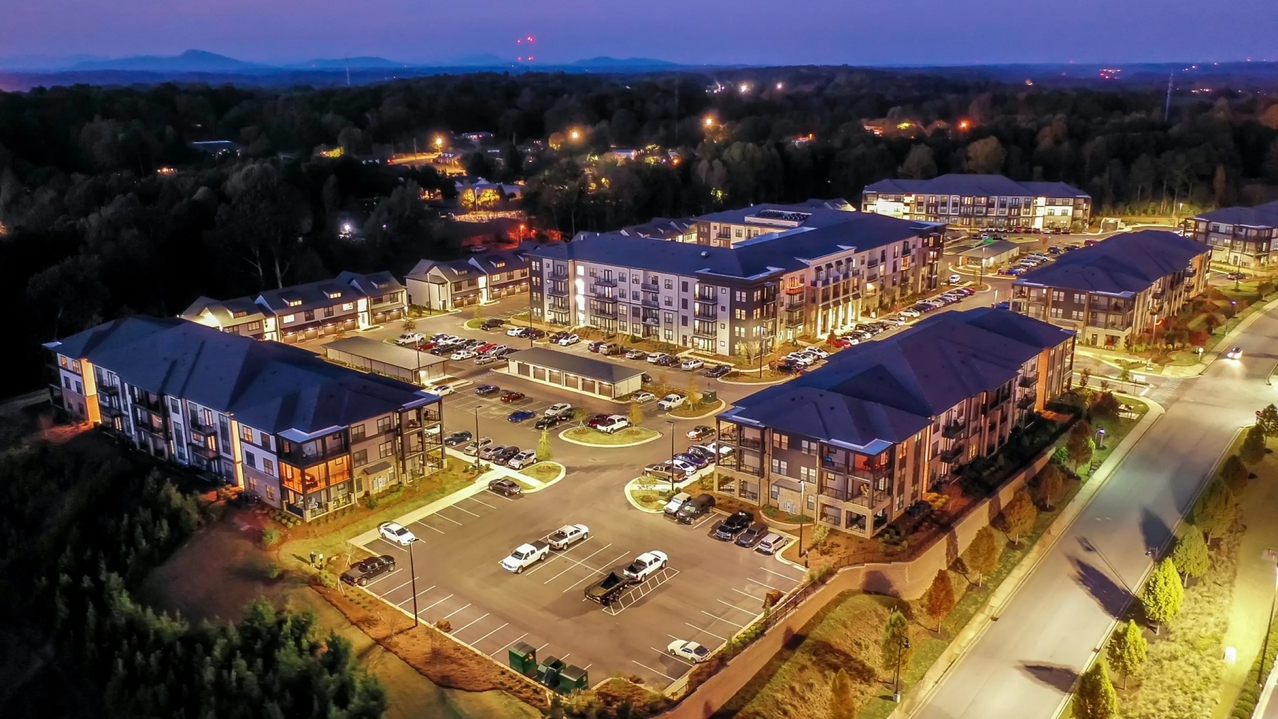 Aerial view of a well-lit residential complex with multiple modern apartment buildings and spacious parking at dusk.