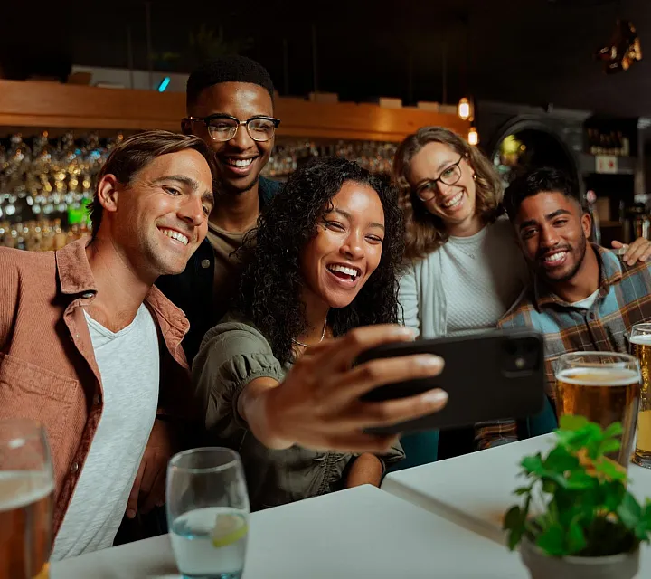 Group of friends taking a selfie at a lively bar, surrounded by drinks and smiling widely.