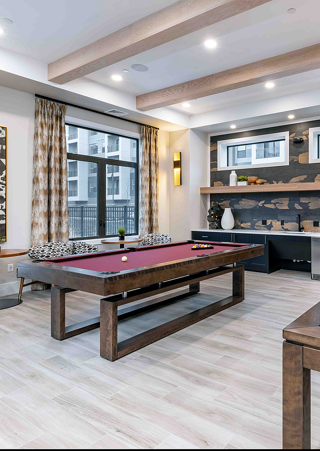 Elegant apartment game room featuring a luxurious pool table, modern shuffleboard, and stylish decor with large windows and cozy seating.