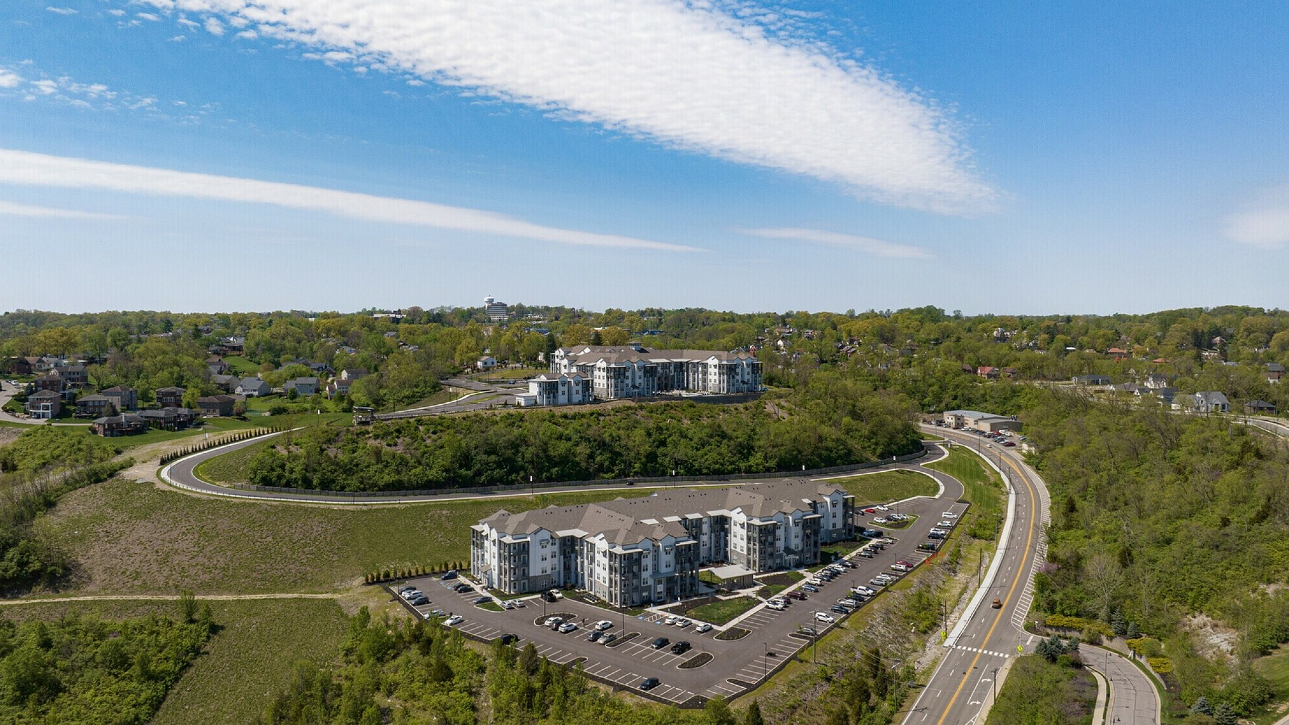 Scenic aerial view of a modern apartment complex nestled in lush greenery with ample parking and winding roads, ideal for serene living.