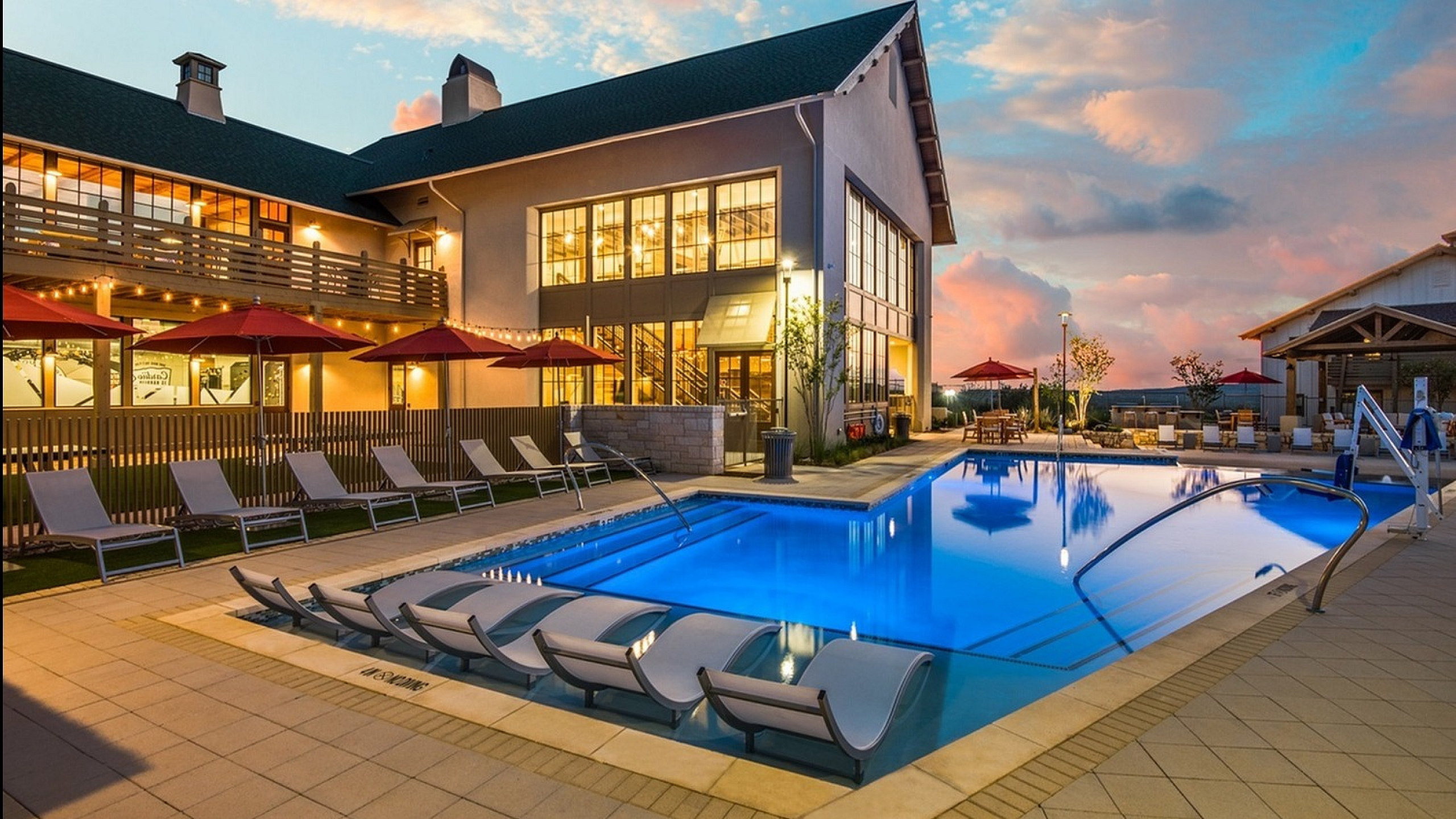 Resort-style pool area with modern lounge chairs, elegant lighting, and a luxurious clubhouse at sunset.