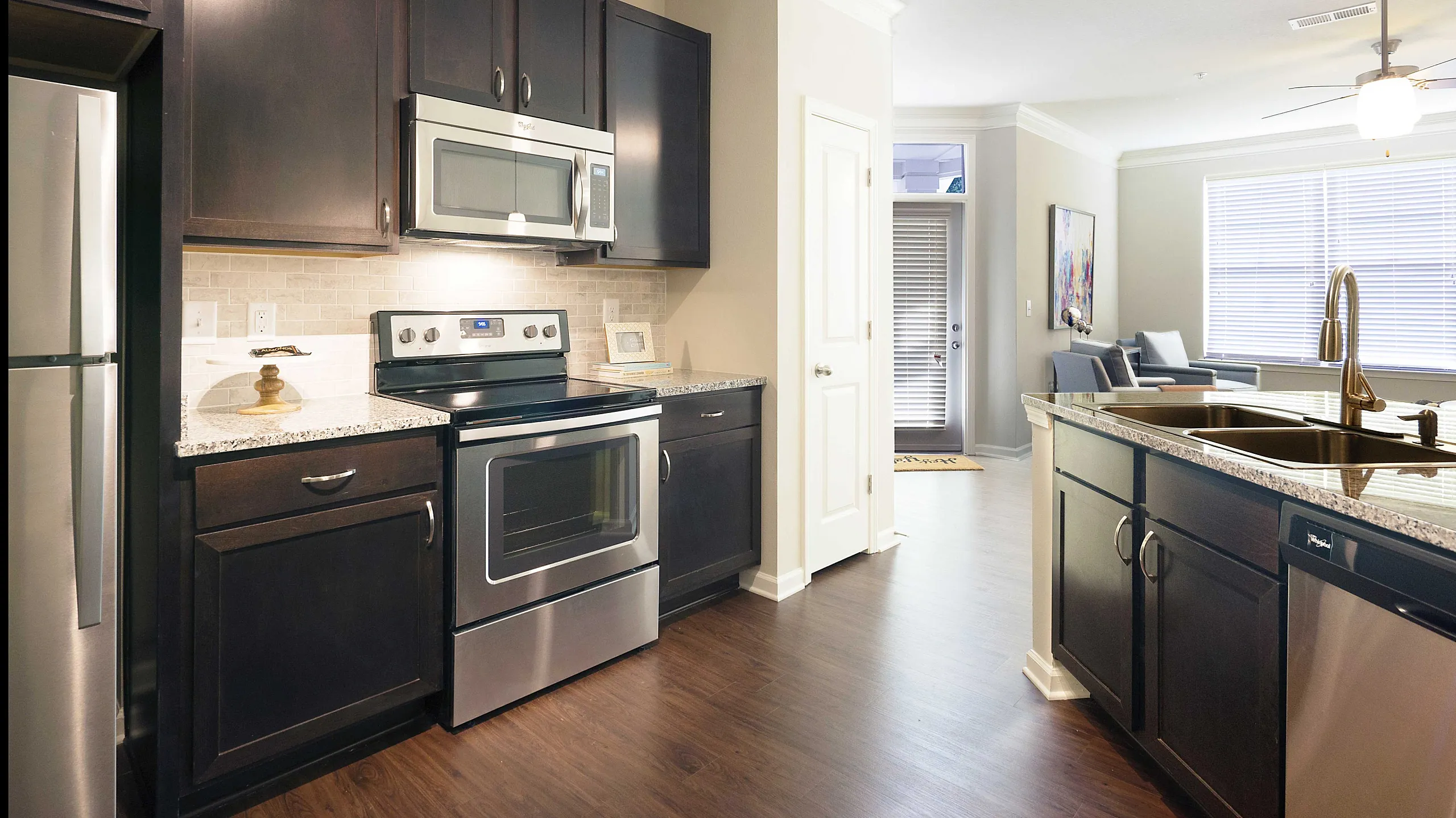 Modern kitchen with sleek stainless steel appliances, dark wood cabinetry, and granite countertops flowing into a bright living space with large windows.