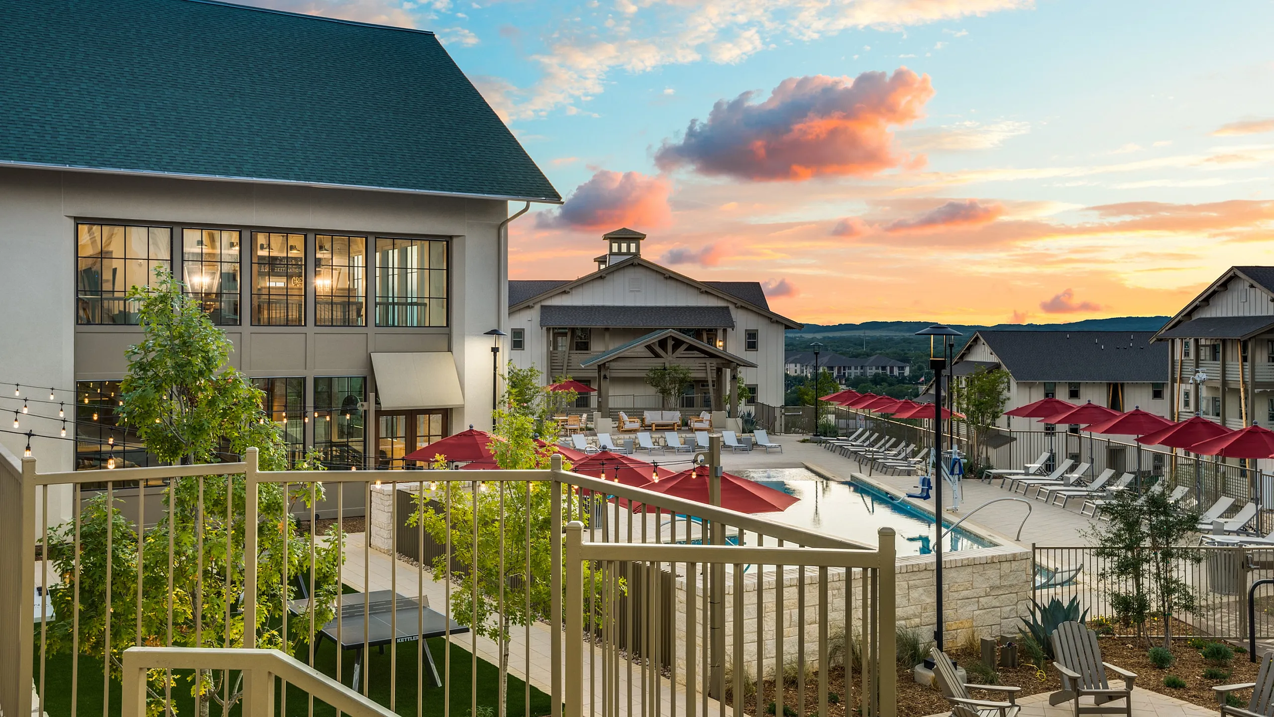 Charming apartment complex with poolside seating, vibrant red umbrellas, and a scenic sunset backdrop, perfect for relaxing and social gatherings.
