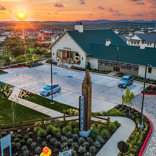 Captivating aerial view of Vecina apartments at sunset, showcasing a modern building with lush landscaping, ample parking, and scenic countryside backdrop.