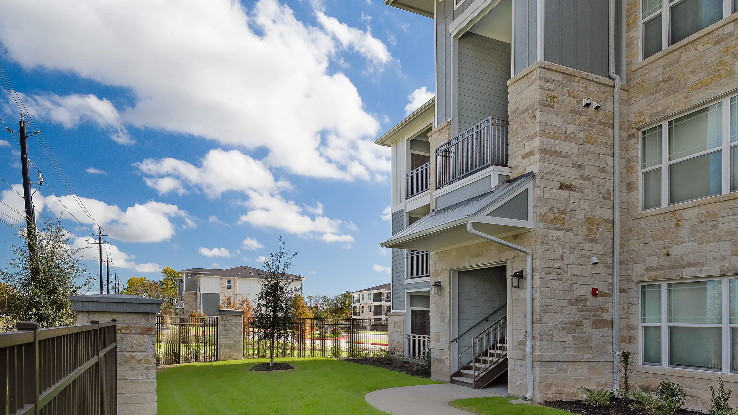 Charming residential building with stone facade, featuring spacious balconies and vibrant green landscaping under a bright blue sky.